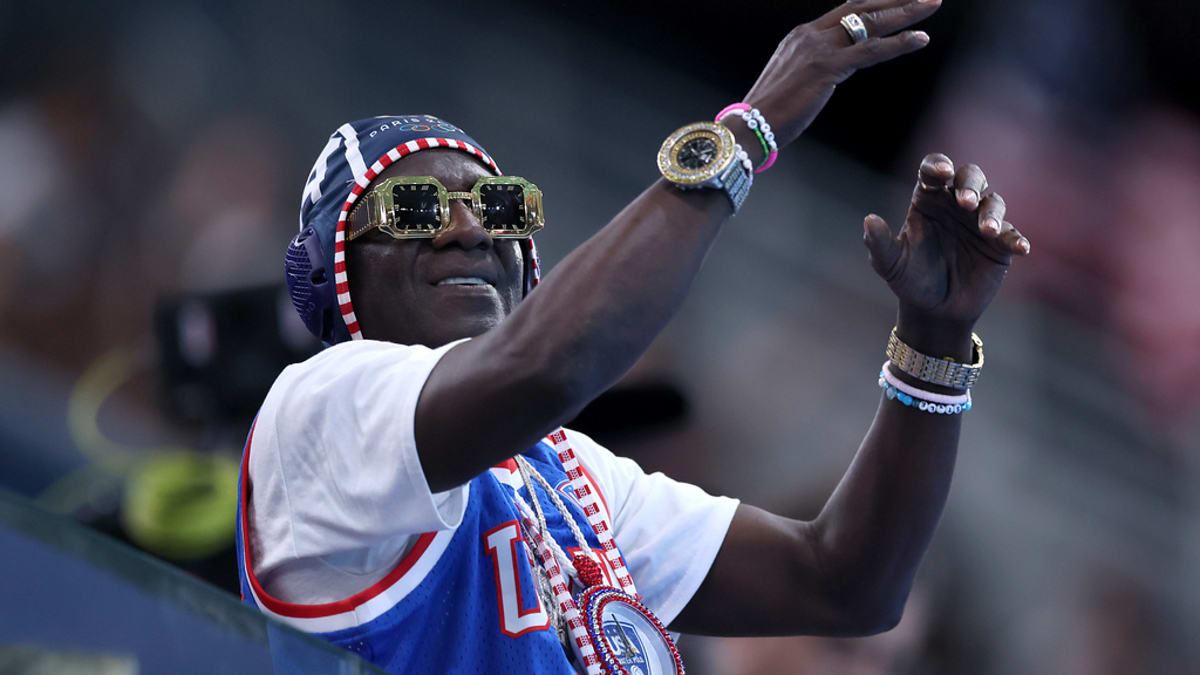 Flavor Flav enthusiastically cheering at a sports event, wearing a USA basketball jersey, large gold clock necklace, and stack of colorful wristbands
