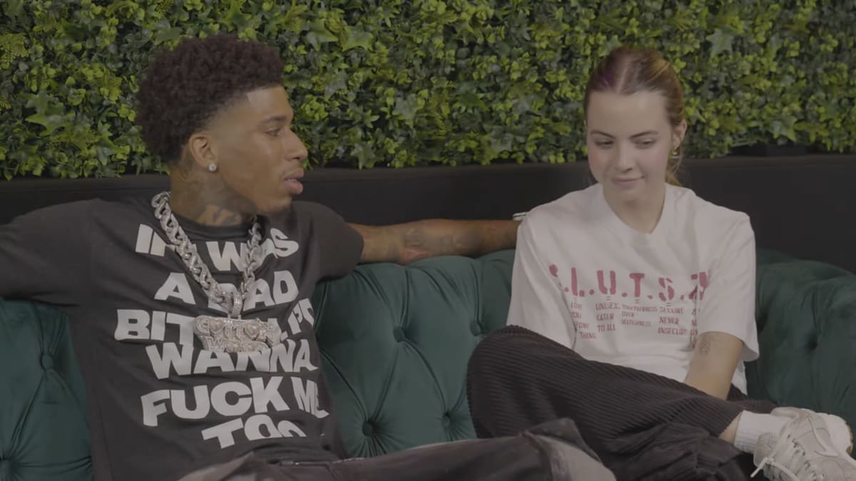 NLE Choppa and AJ Clementine sit on a sofa in a relaxed setting. NLE Choppa is wearing a shirt with text and large chains; AJ is in casual wear