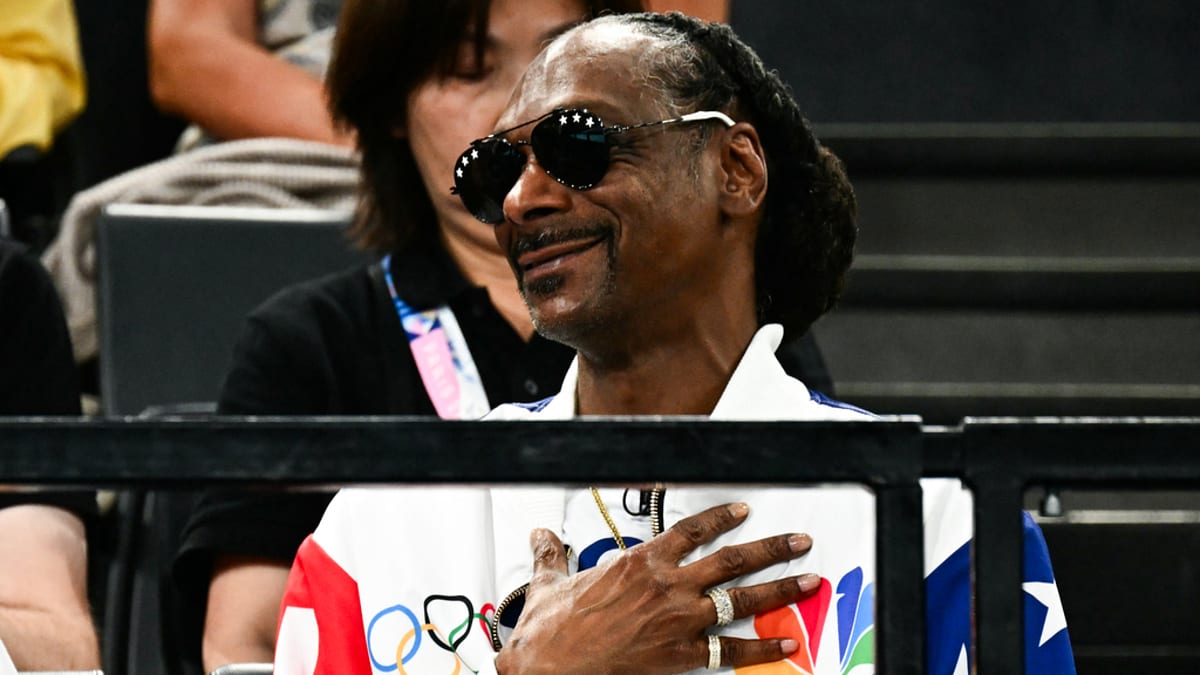 Snoop Dogg at an event, wearing sunglasses and an Olympic-themed jacket with his hand on his chest, smiling