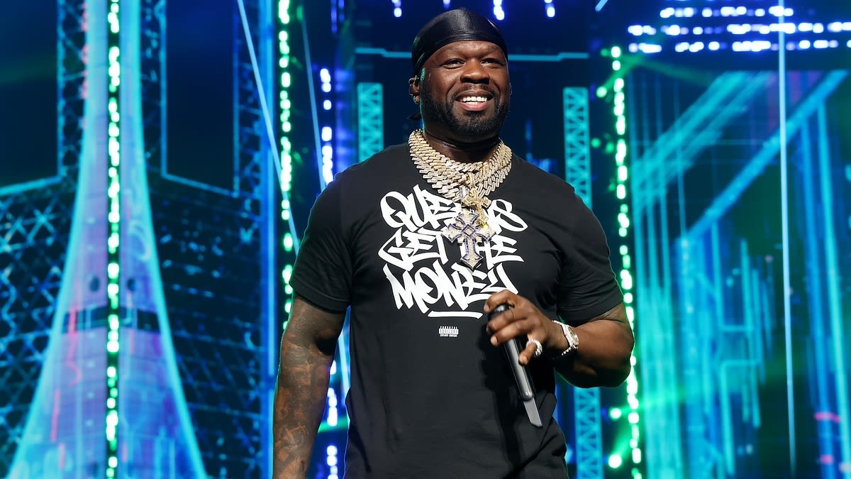 50 Cent performs on stage wearing a black T-shirt with graffiti-style text, layered gold chains, and a black bandana. High-energy background lighting