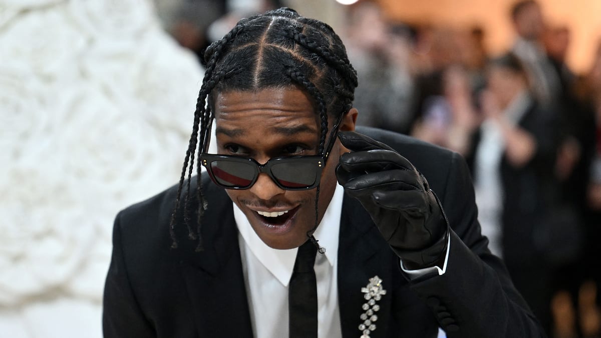 A$AP Rocky in a black suit and tie, wearing sunglasses and a flower brooch, smiles and looks over his glasses