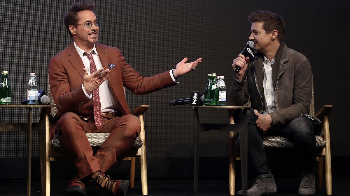 Robert Downey Jr. in a suit and Jeremy Renner in a casual jacket are seated on stage, engaging in conversation at a media event