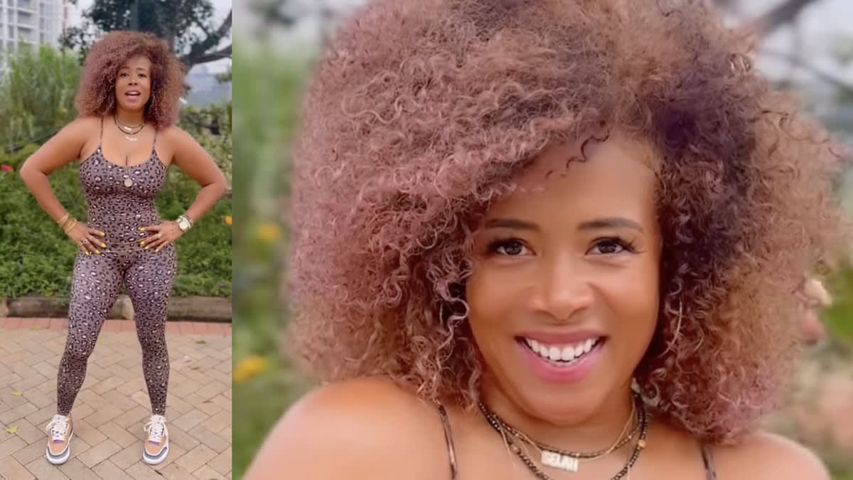 Kelis wears a leopard-print jumpsuit and sneakers, with gold necklaces and voluminous curly hair. She stands outdoors and smiles brightly in the close-up shot