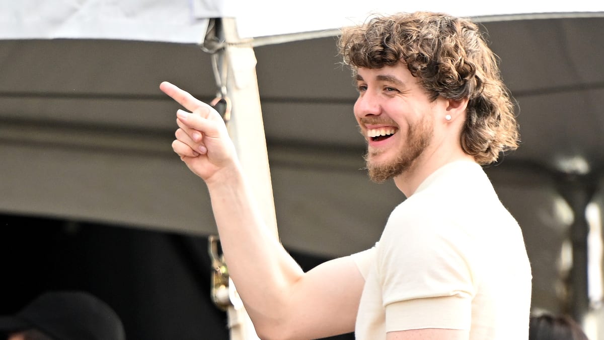 Jack Harlow smiling and pointing during an outdoor event. He is dressed casually with a light shirt