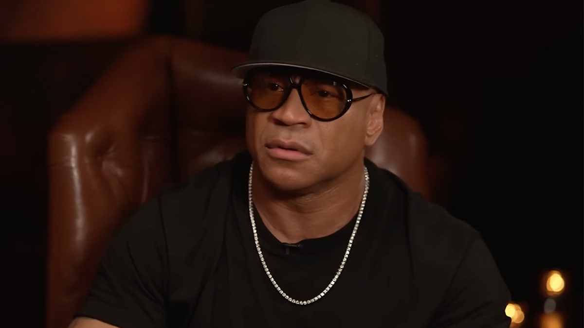 LL Cool J in a black T-shirt and chain necklace, seated in a brown leather chair, wearing glasses and a black baseball cap