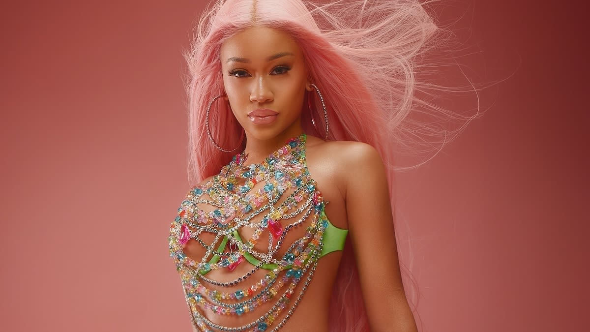 Saweetie poses in a vibrant outfit adorned with bejeweled chains and accessories against a plain background