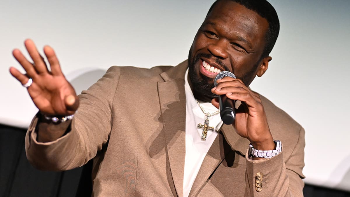 50 Cent is smiling, holding a microphone, and gesturing with his hand while wearing a suit jacket and a cross necklace