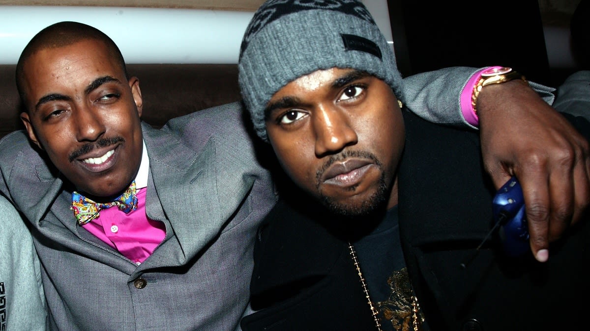 Enyce Smith and musician Kanye West are posing together. Kanye West is wearing a beanie and dark jacket, while Enyce Smith wears a suit with a colorful bow tie