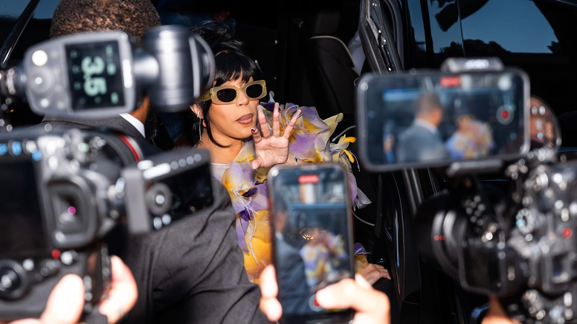 Cardi waves to cameras while stepping out of a car in an extravagant, feathered outfit amid flashing lights and photographers