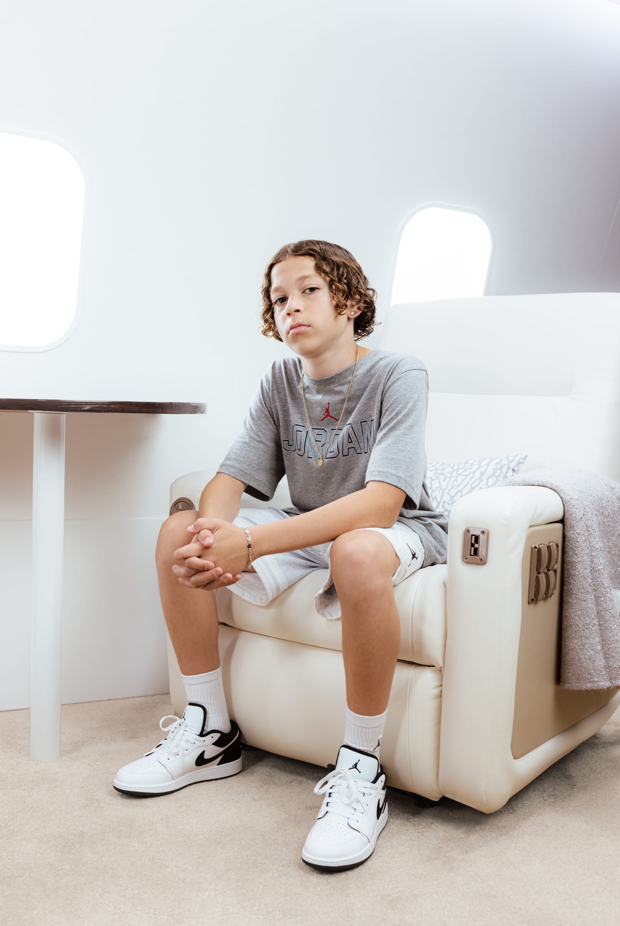 A young person with curly hair sits in a luxurious airplane seat, wearing a gray Jordan shirt, shorts, and white Nike sneakers.