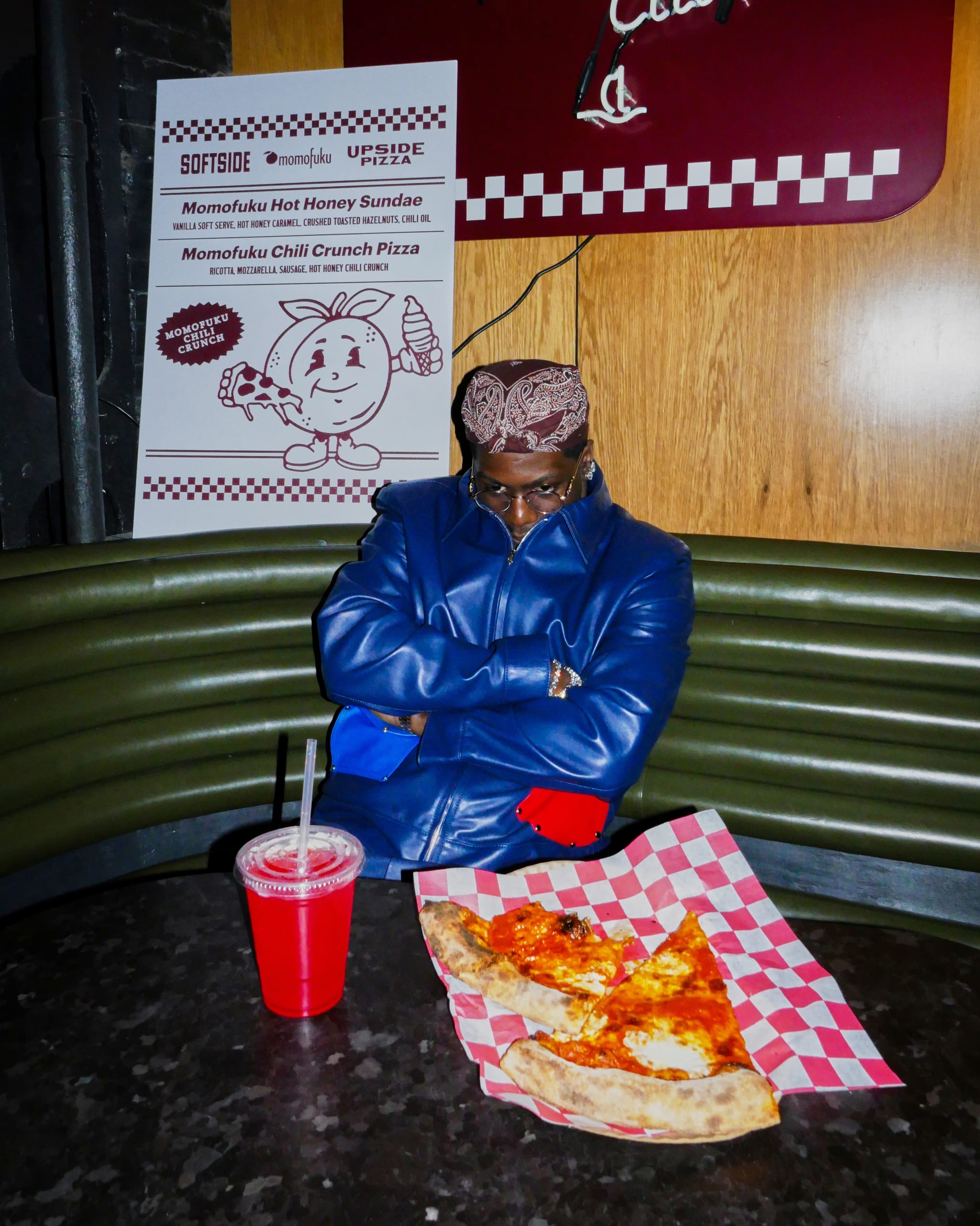 A person in a blue jacket and bandana sits at a table with pizza slices and a red drink. A sign behind advertises Momofuku items.