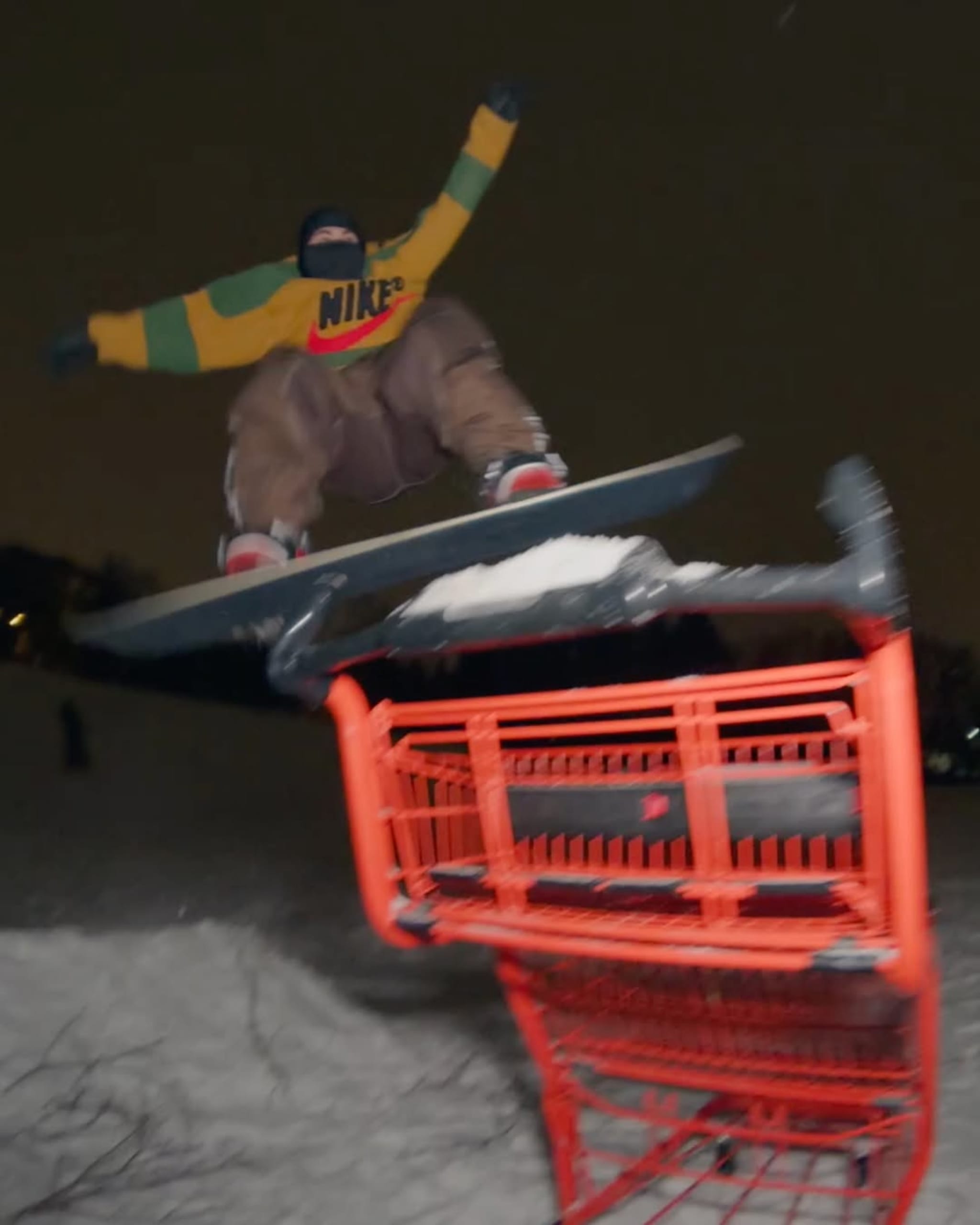 A snowboarder in a Nike jacket performs a jump at night, with a blurred background. Nike x Cactus Plant Flea Market
