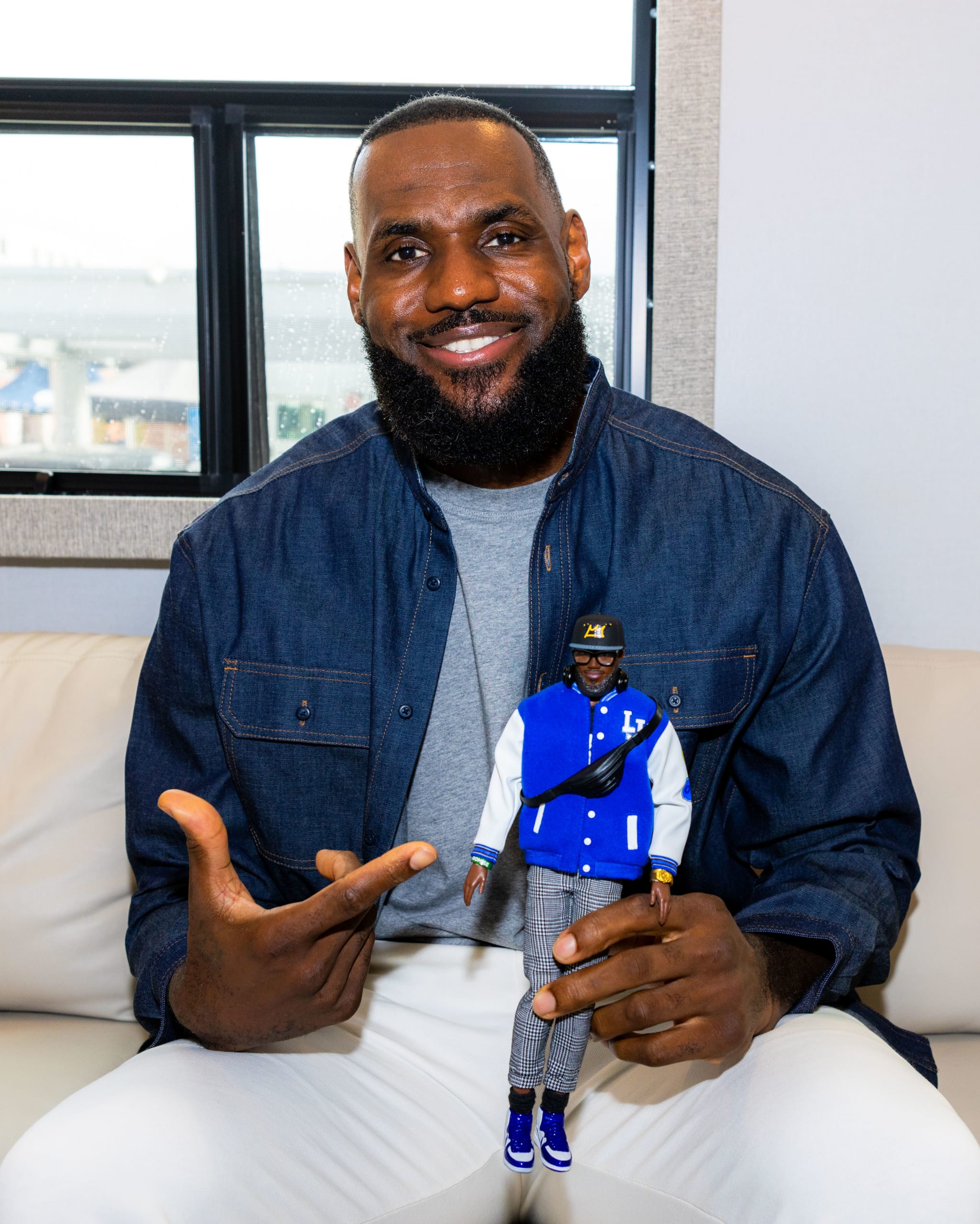 LeBron James smiling, seated, holding a small action figure of himself, wearing a denim jacket and light pants.