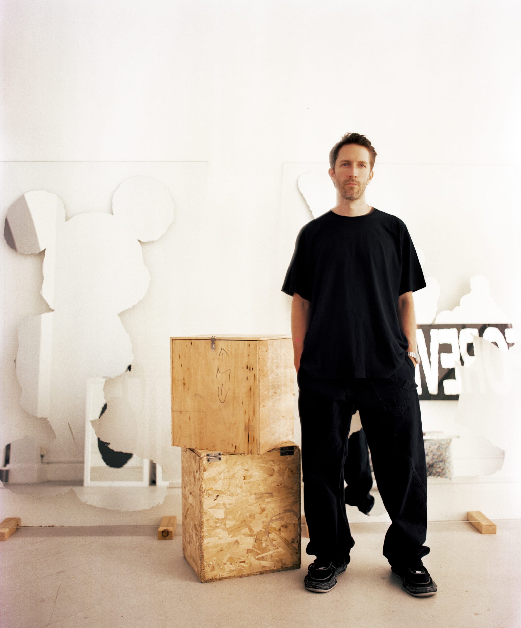 A man in black clothing stands beside wooden boxes, with abstract art pieces in the background.