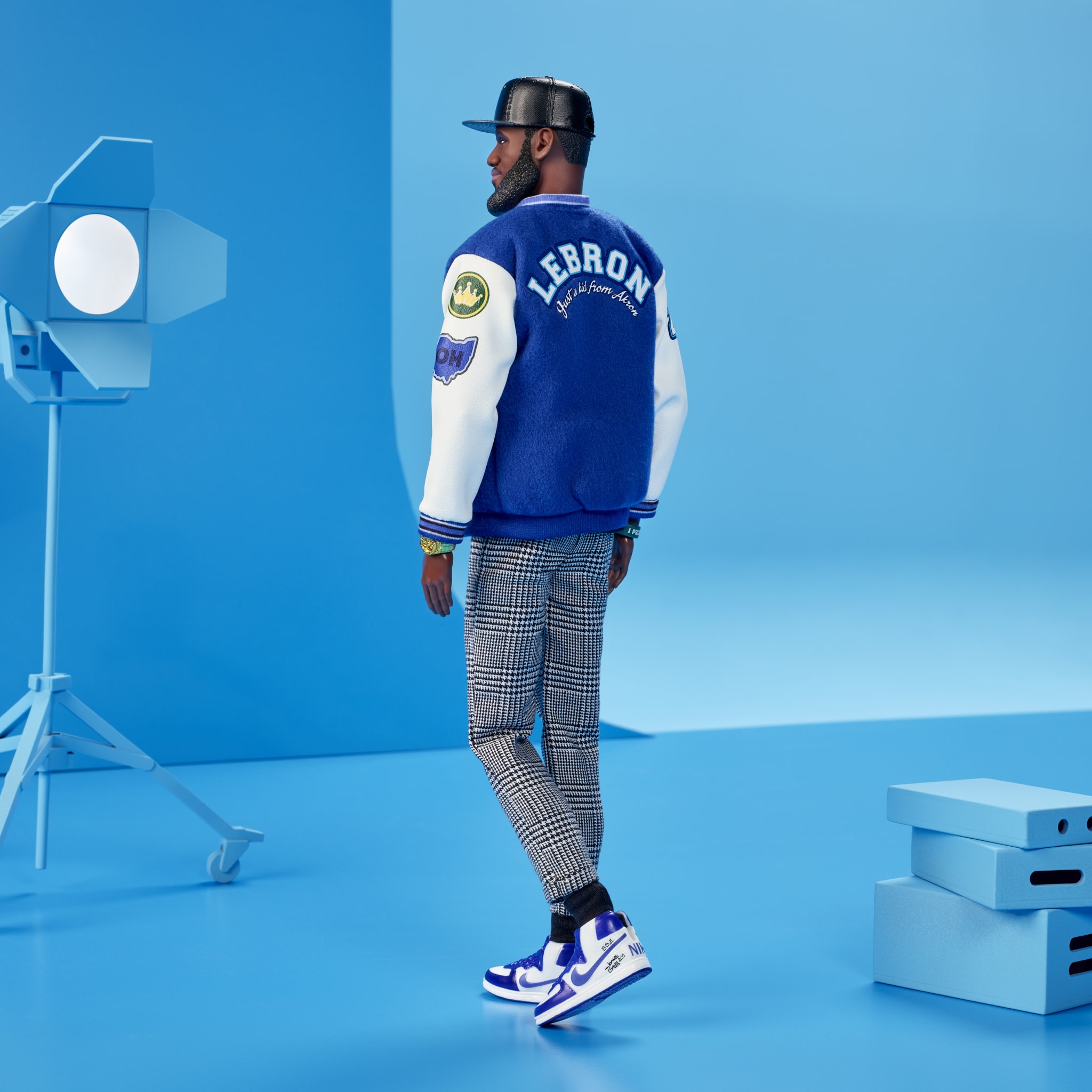 A figure wearing a blue and white "LeBron" jacket, plaid pants, and sneakers stands in a blue studio setting with a spotlight.