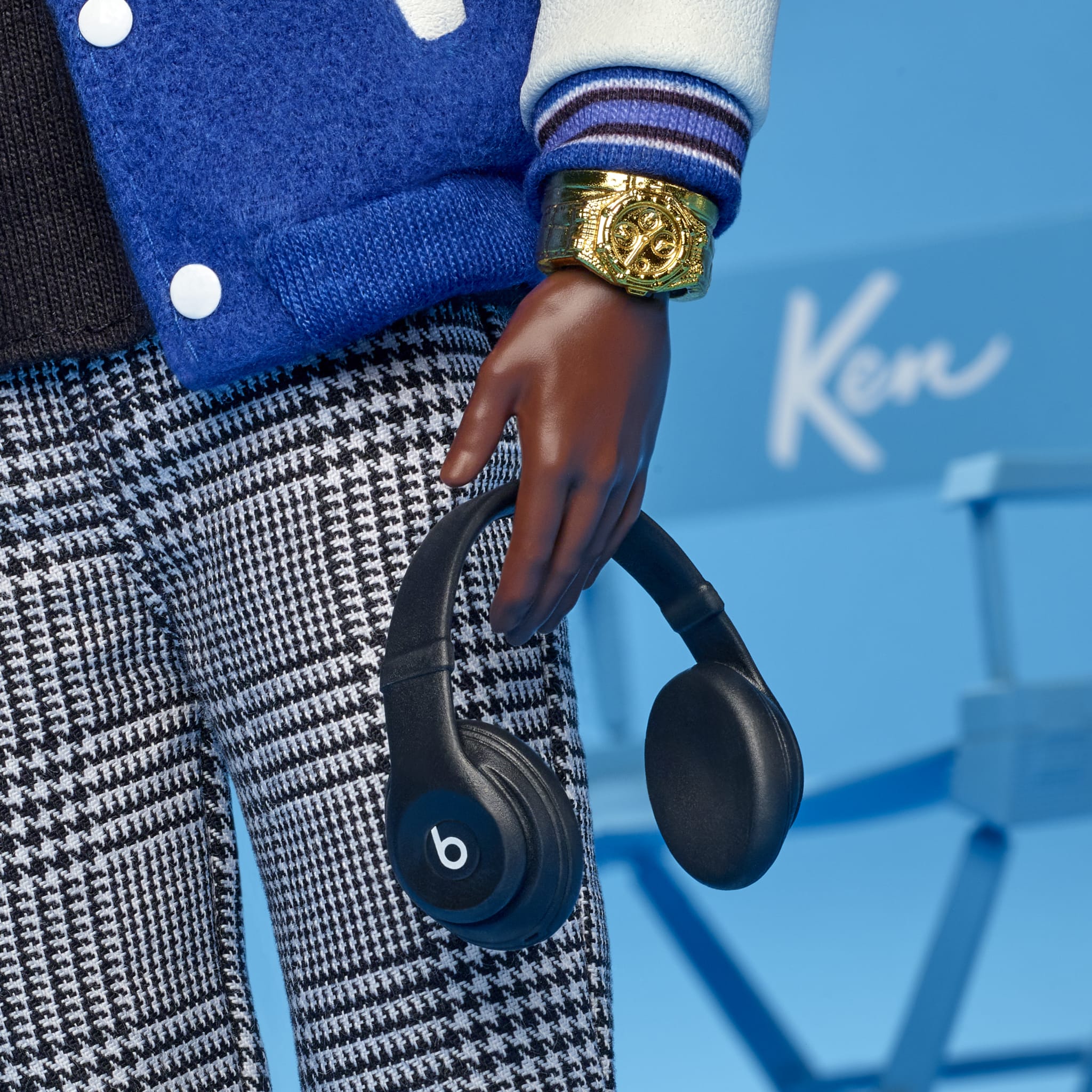 A doll's hand holding black Beats headphones, wearing a gold watch, blue varsity jacket, and checkered pants. "Ken" is visible in the background.