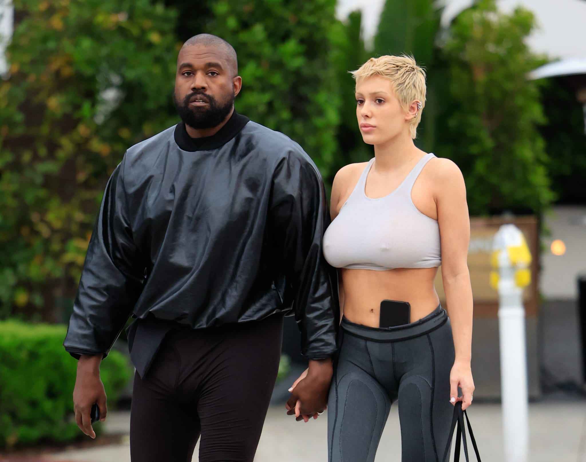 Kanye West, in a black outfit, walks hand in hand with Bianca Censori, wearing a grey crop top and leggings, outdoors.