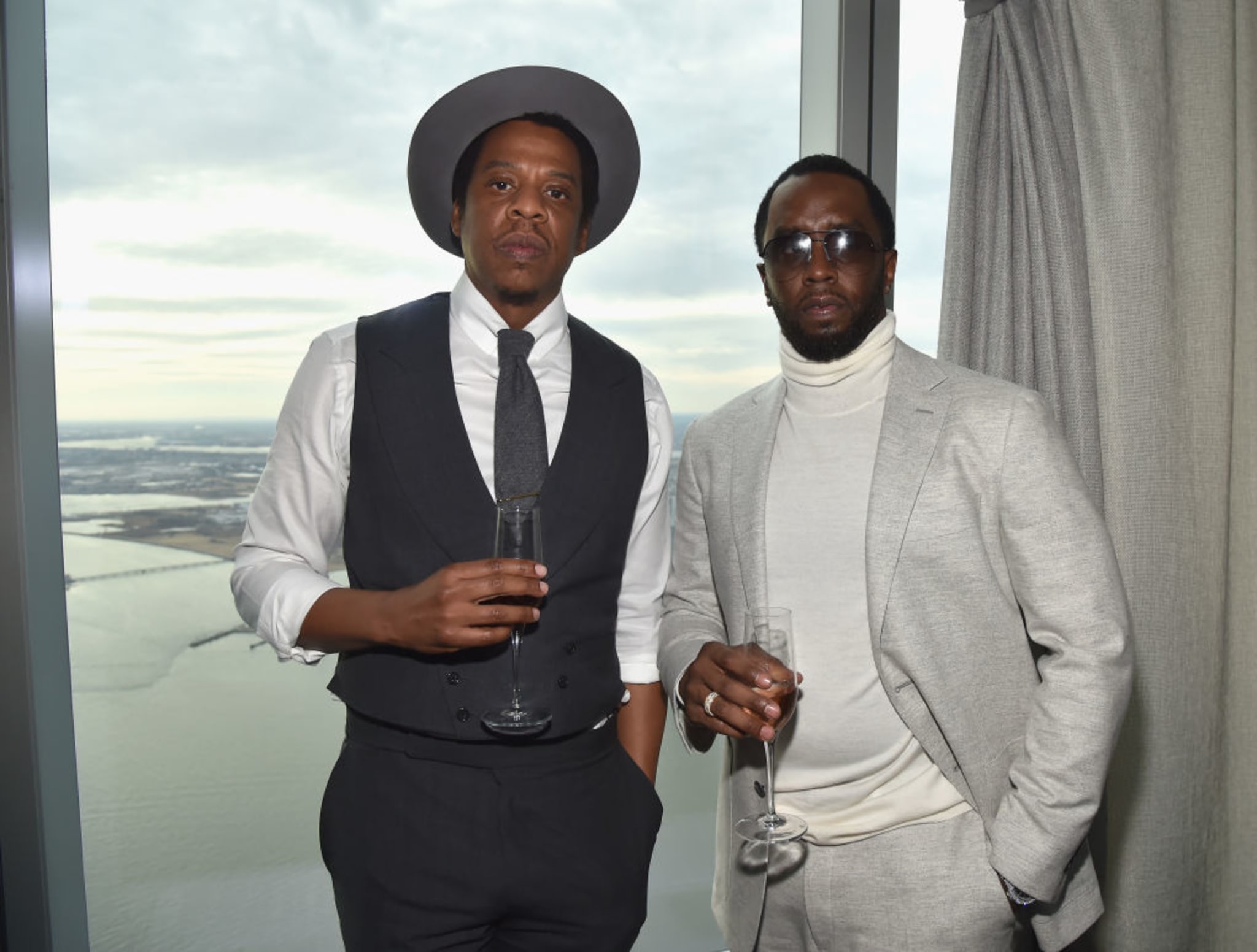 Jay-Z and Diddy stand by a window holding champagne glasses. Jay-Z wears a hat and vest, while Diddy is in a light gray suit.