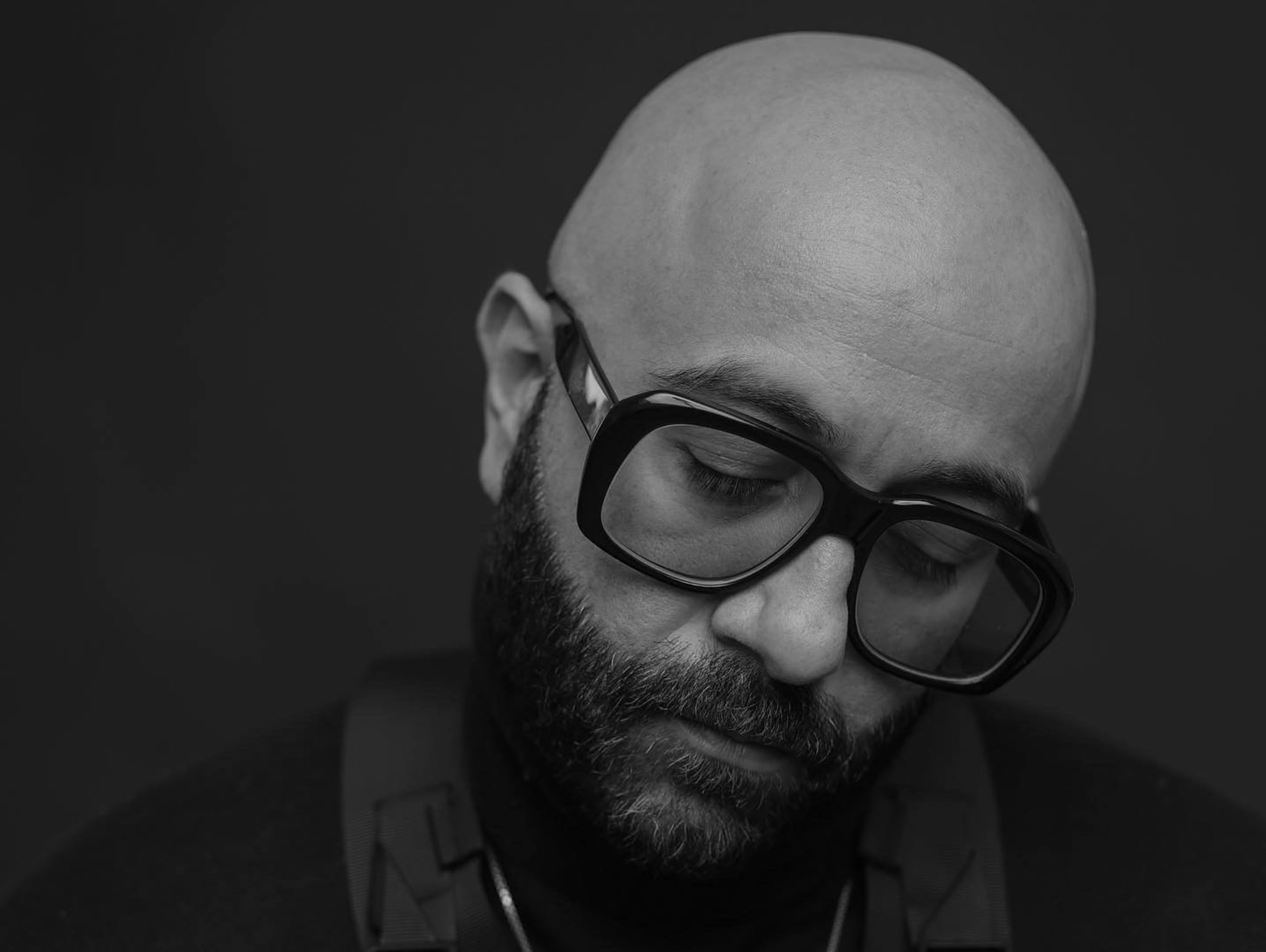Black and white image of Narcy looking down. He is wearing thick rimmed glasses and a chain