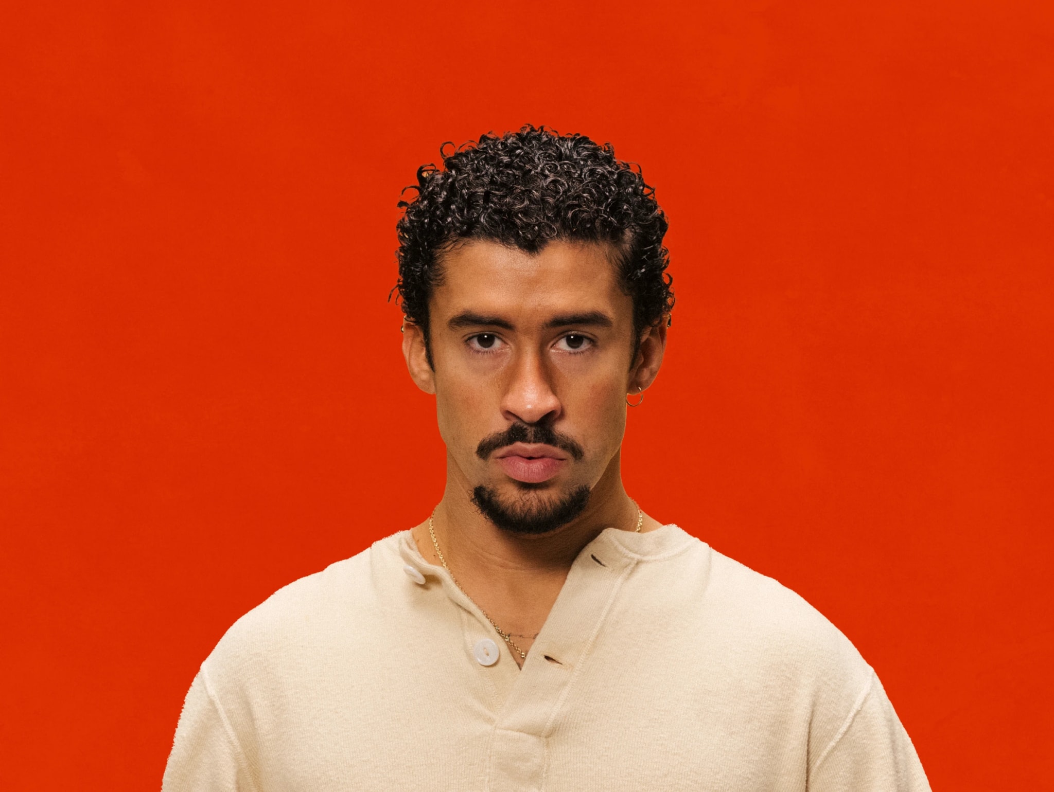 A man with curly hair and a goatee, wearing a beige shirt, stands against an orange background.