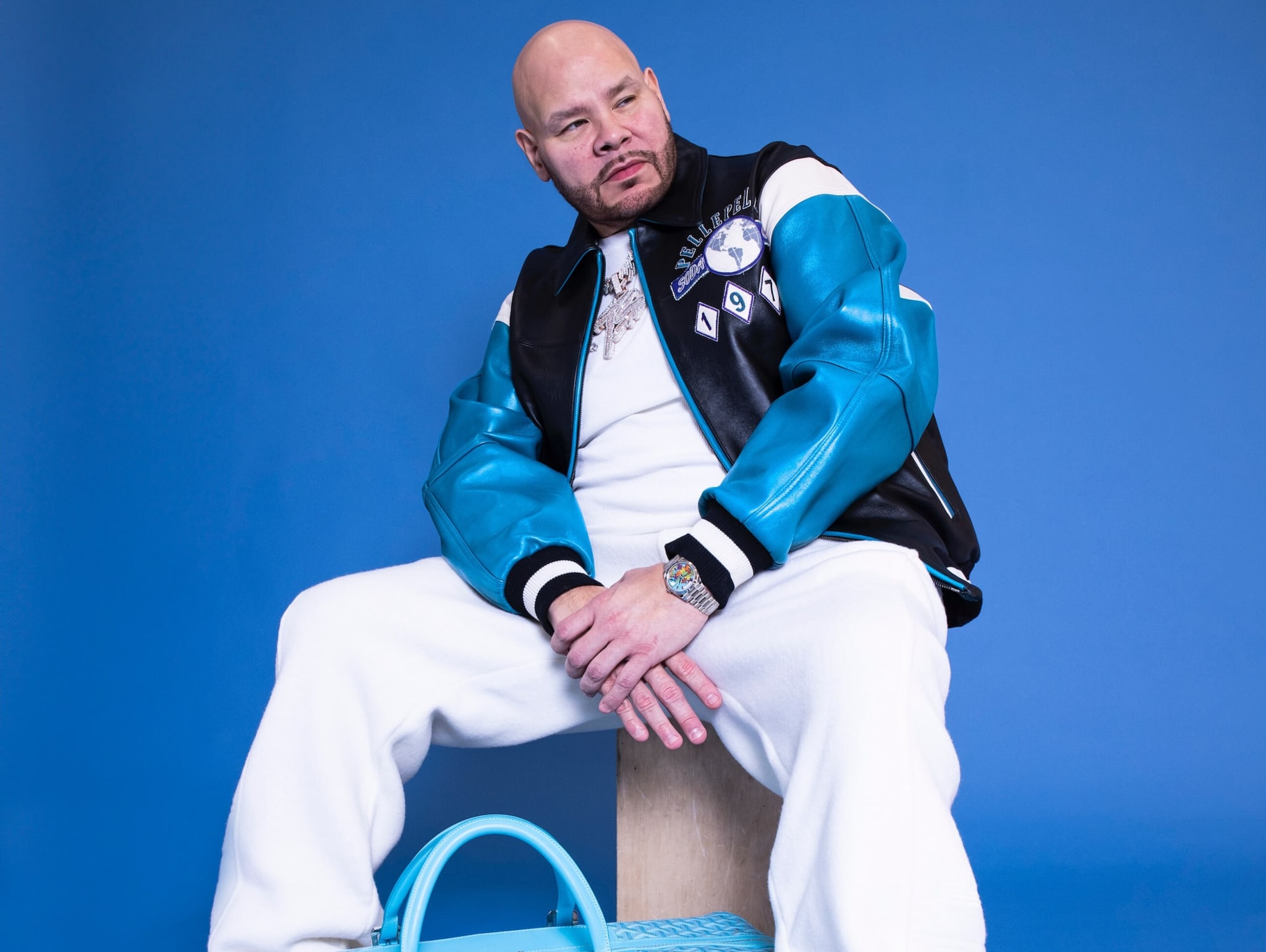 Fat Joe sitting against a blue background, wearing a blue and black jacket, white pants, and a watch, with a blue bag nearby.