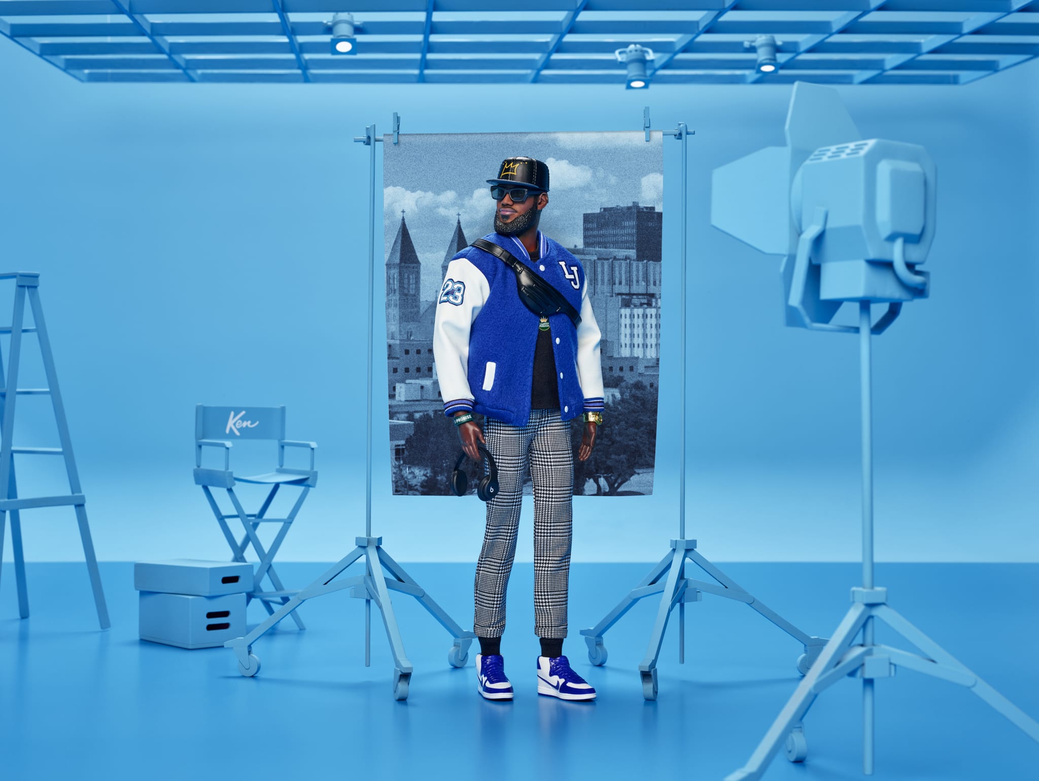 A stylish man in a blue varsity jacket and plaid pants stands in a blue-toned studio with photography equipment.