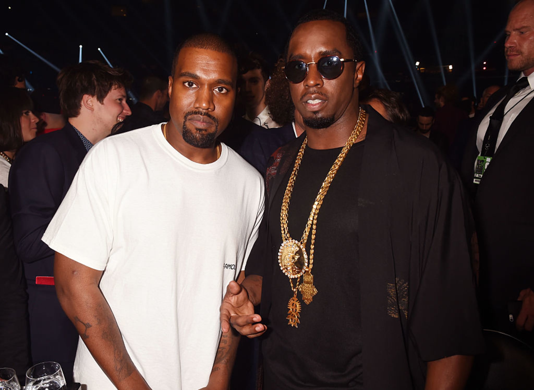 Kanye West and Sean "Diddy" Combs at an event, with Kanye in a white t-shirt and Diddy wearing sunglasses and a black outfit with gold chains.