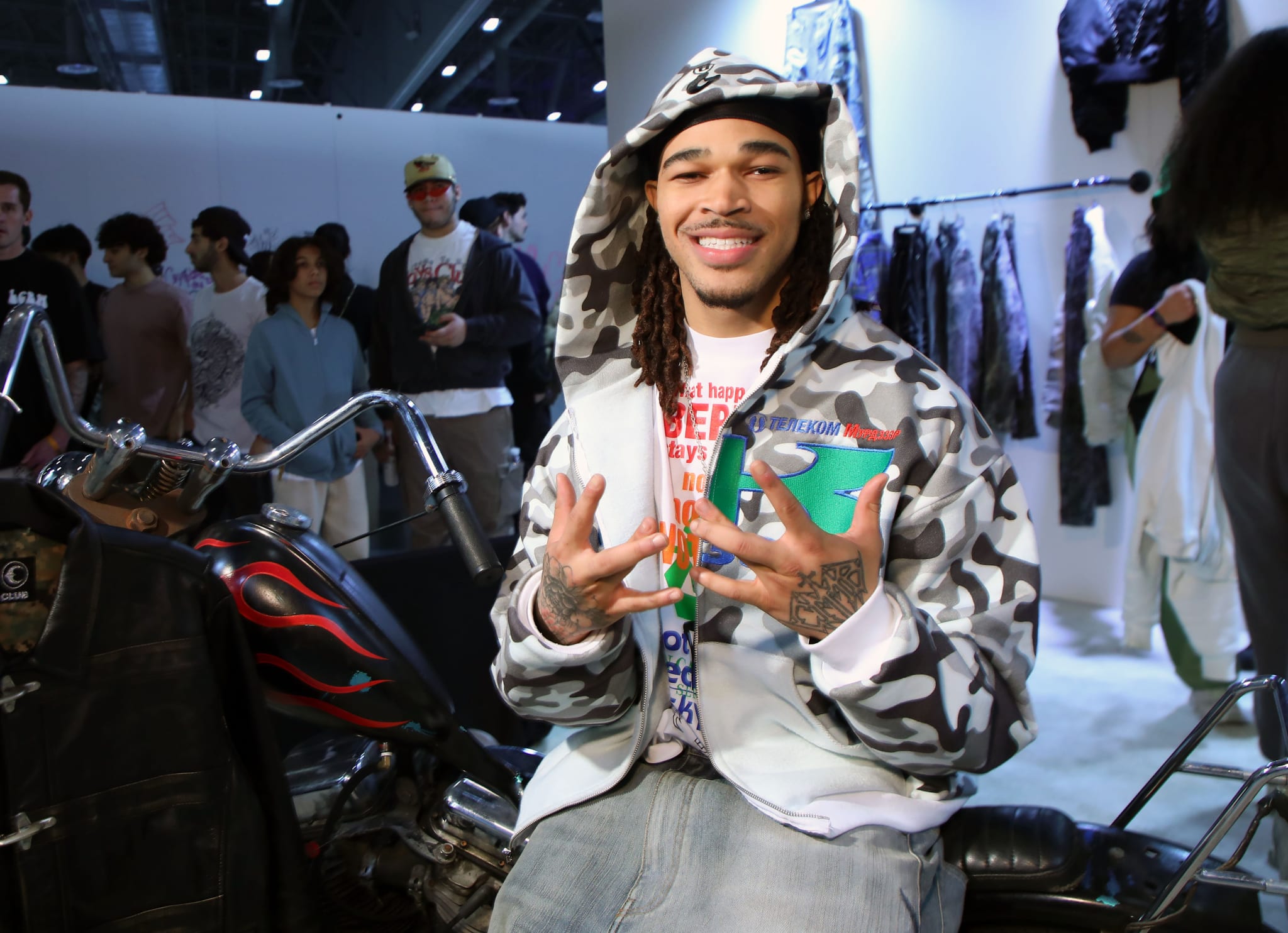 Plaqueboymax sits on a motorcycle indoors, smiling and gesturing with his hands. People and clothing racks are in the background.