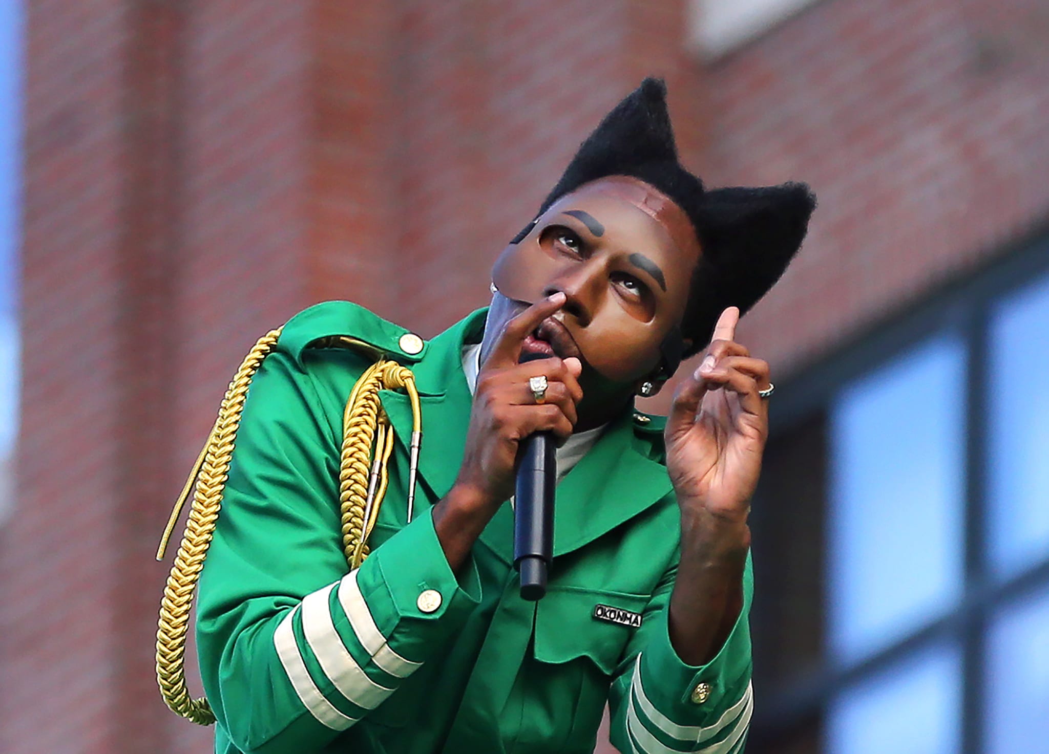 Tyler, the Creator in a green outfit and unique mask sings passionately, holding a microphone, with a brick building in the background.
