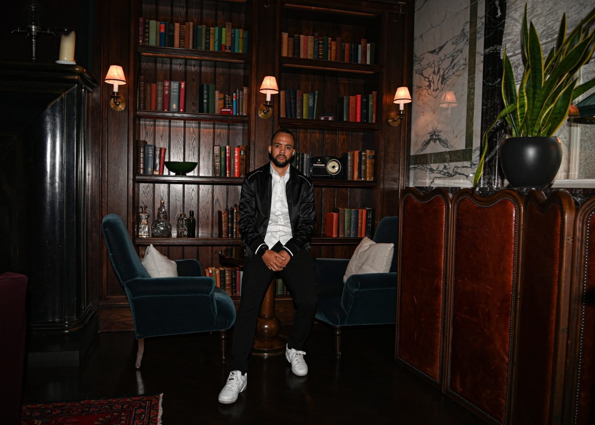 A man in a casual outfit consisting of a white shirt, black jacket, black pants, and white sneakers is seated in a cozy library setting with bookshelves and armchairs