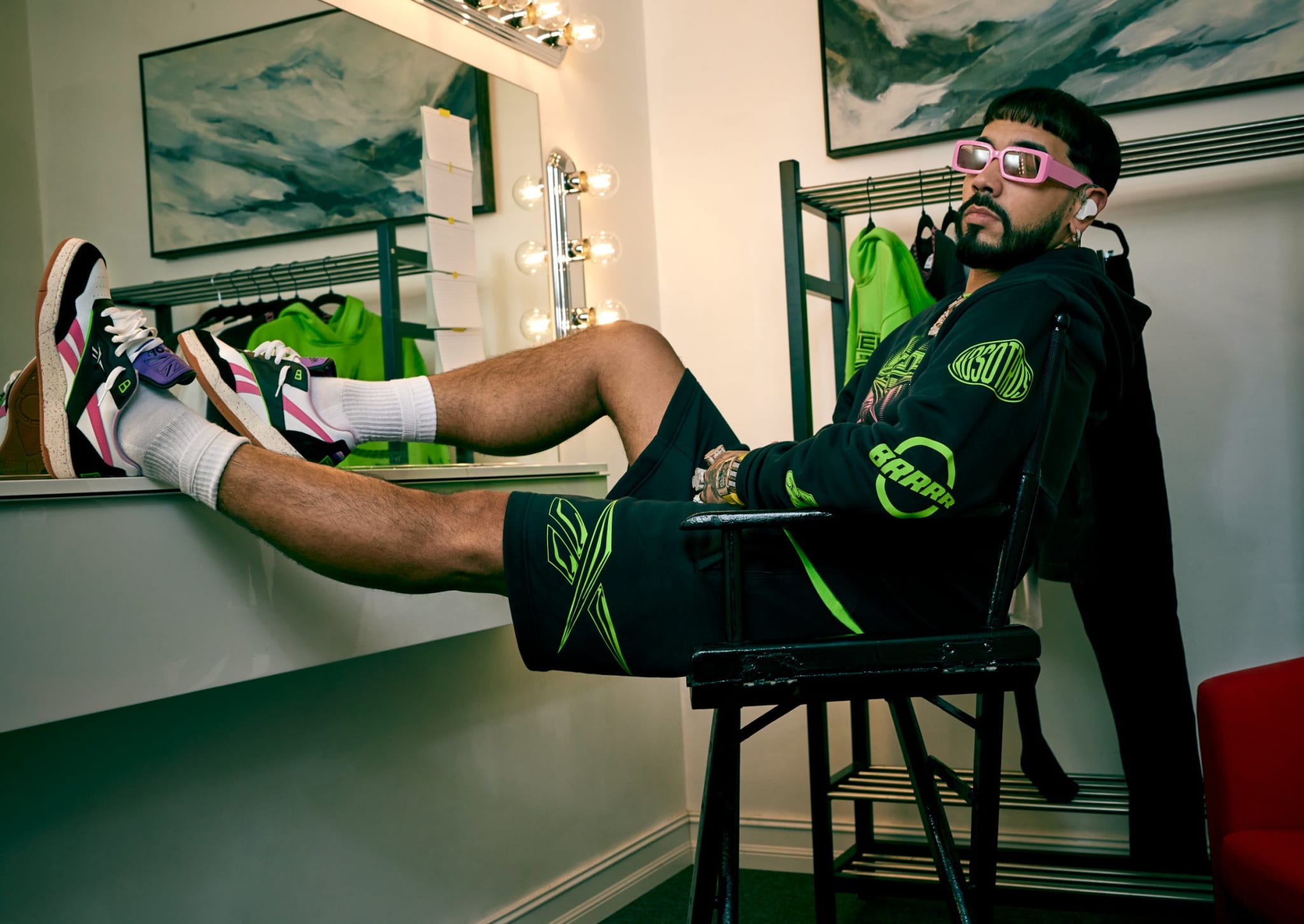 A person lounges in a dressing room, wearing sporty sneakers, athletic shorts, a hoodie, and sunglasses