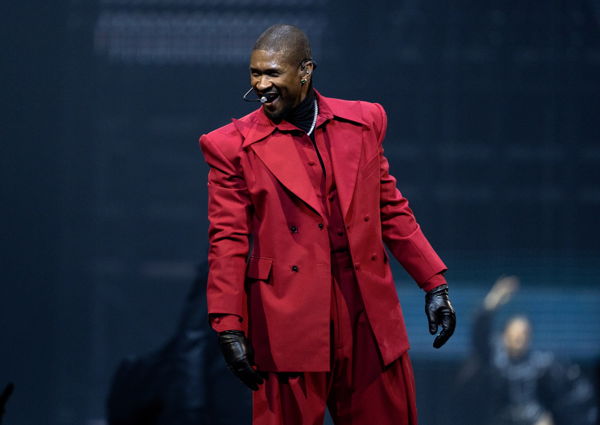 Usher in a red suit and black gloves is on stage with a microphone headset, smiling and engaging with the audience.