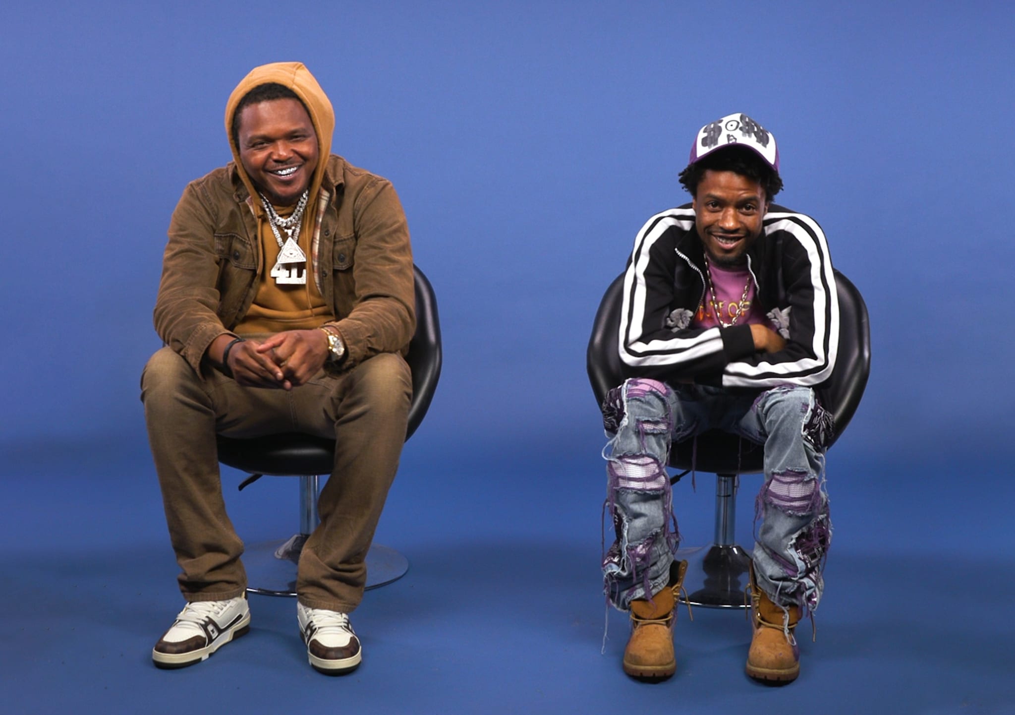Young Nudy and Pi’erre Bourne sitting on chairs against a blue background. Judy wears a brown hoodie, Pi’erre has a black jacket with a cap and ripped jeans.