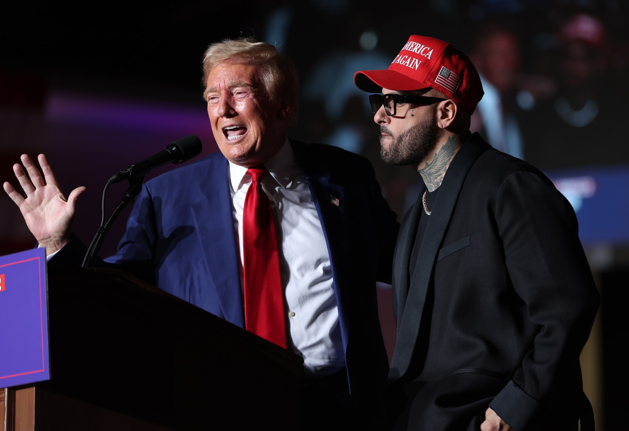 Donald Trump speaking at a podium, wearing a suit and red tie, next to Nicky Jam in a "Make America Great Again" hat and sunglasses.