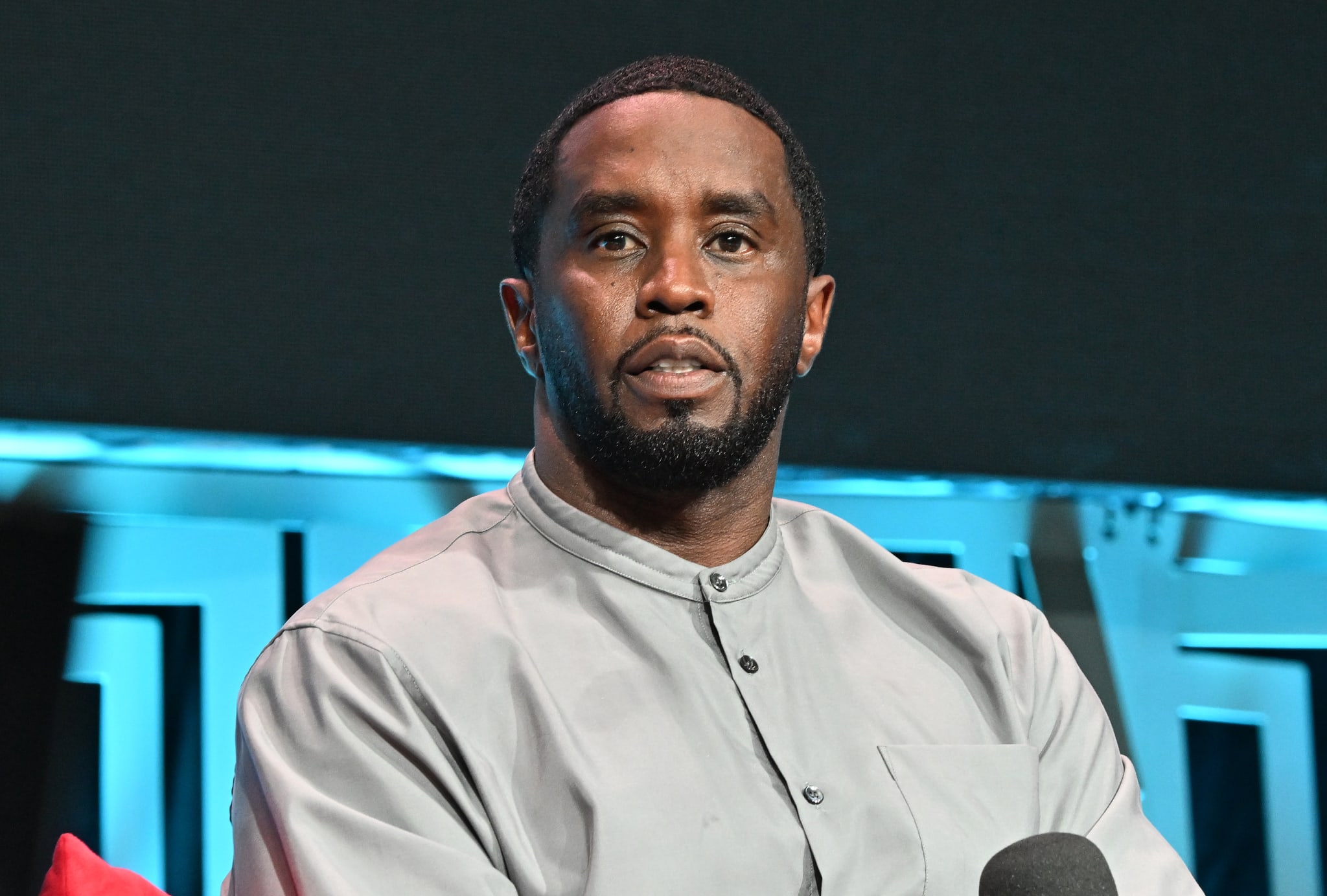 Sean "Diddy" Combs in a light gray shirt, sitting and looking forward, with a microphone in front of him.