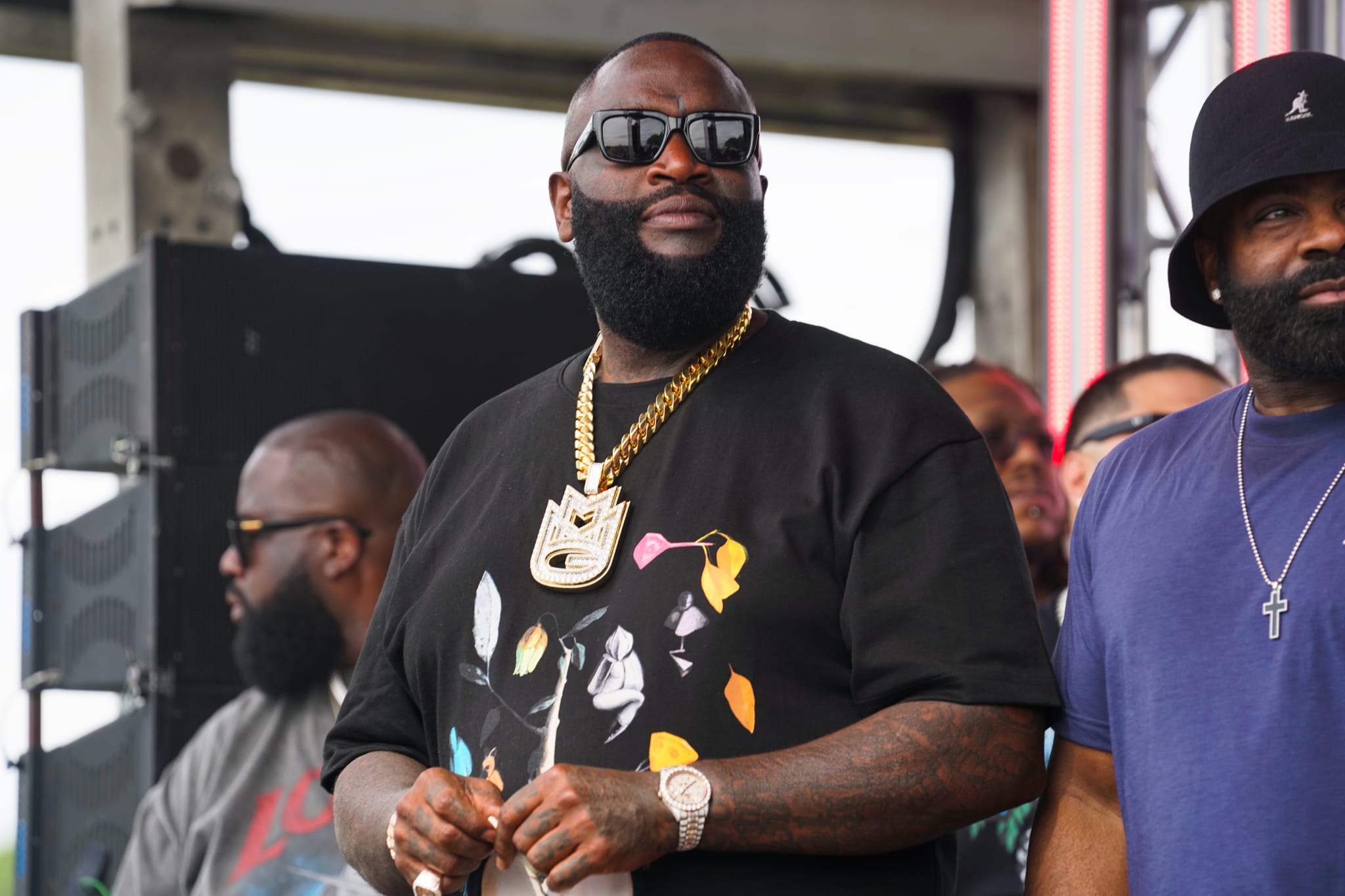 Rick Ross in a black t-shirt with a gold chain and sunglasses, attending a sneaker event