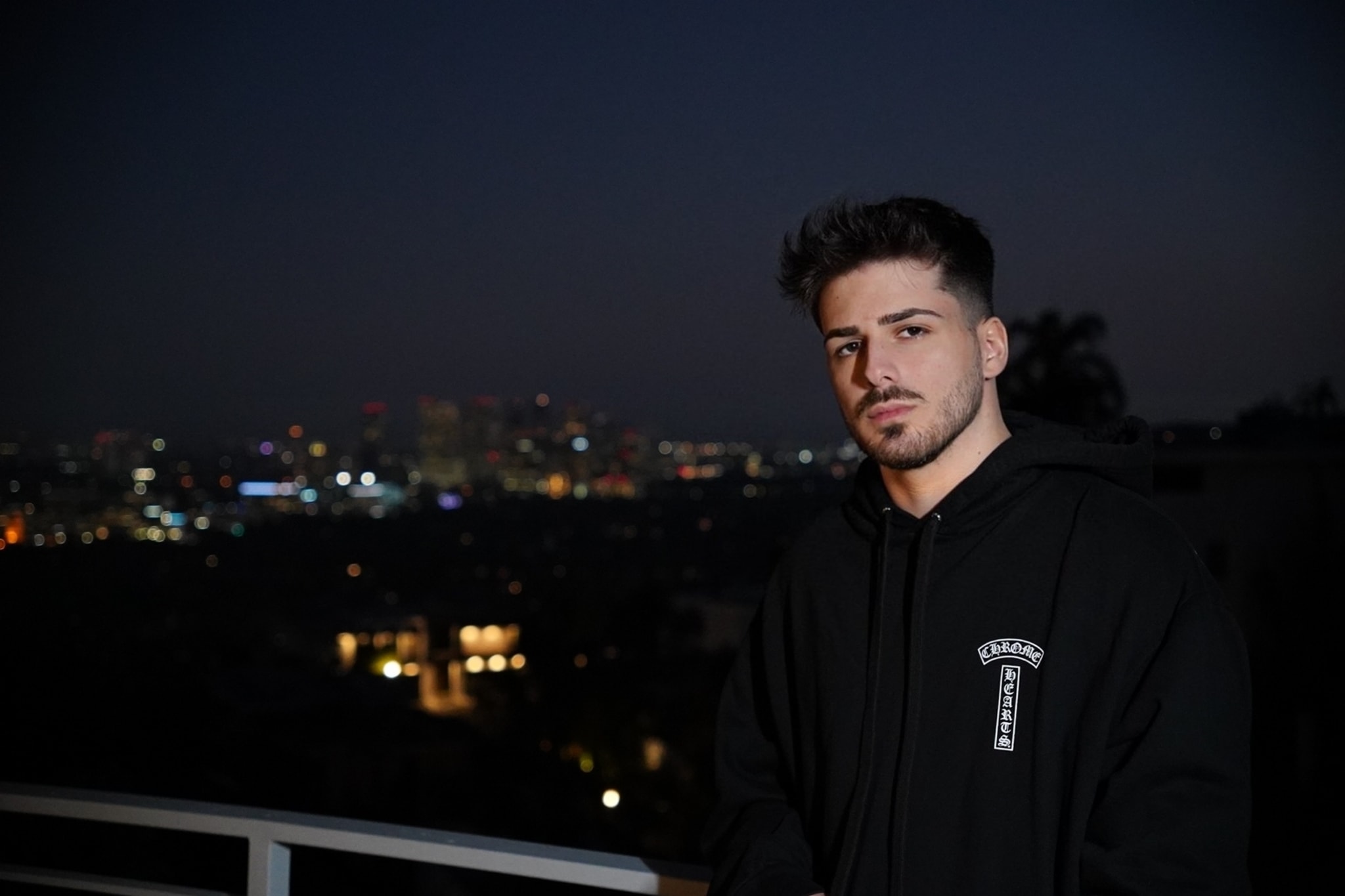 A person stands on a balcony at night with city lights in the background. They are wearing a hoodie and have a serious expression
