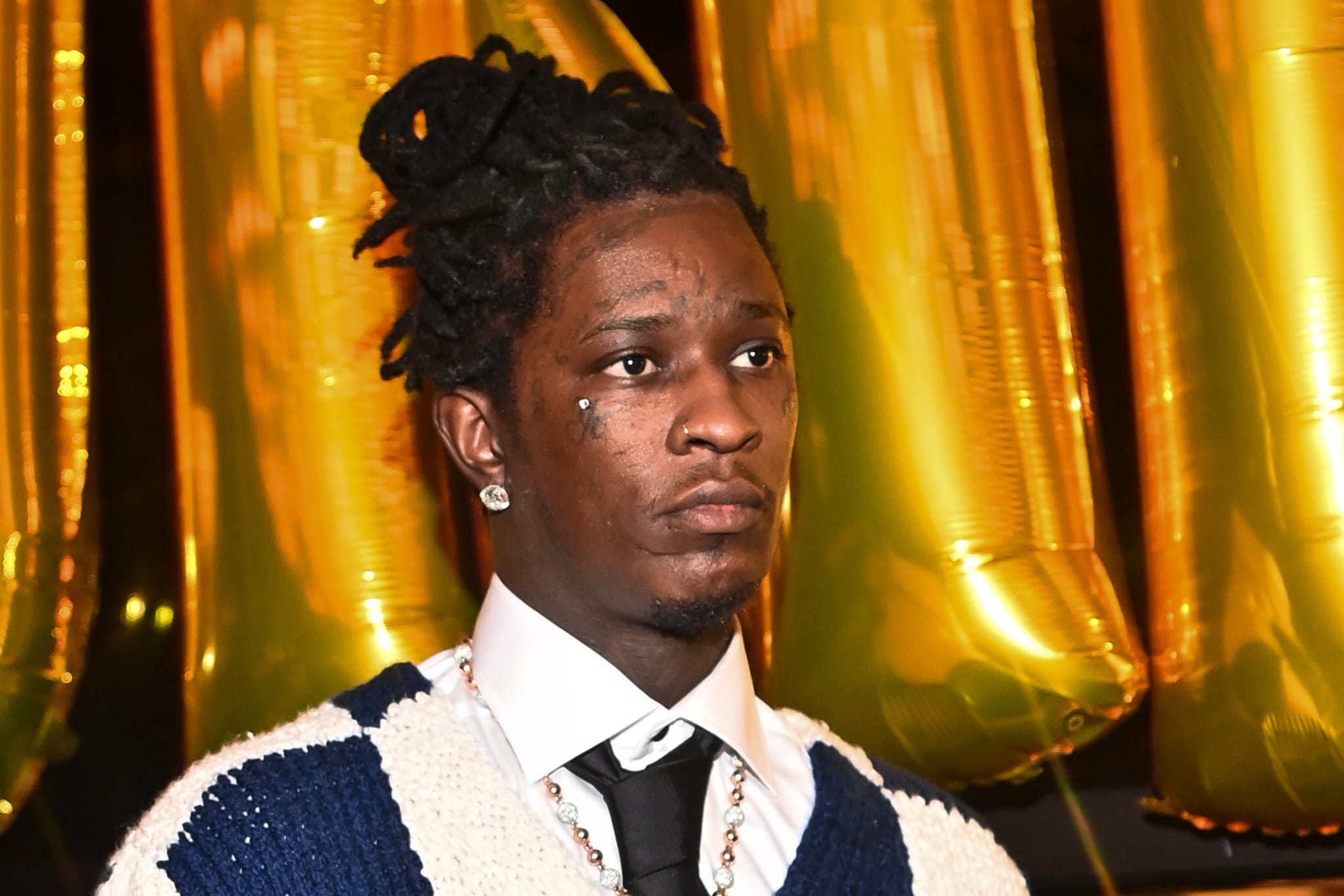 Young Thug wearing a white shirt, black tie, and knitted sweater, standing in front of gold balloons