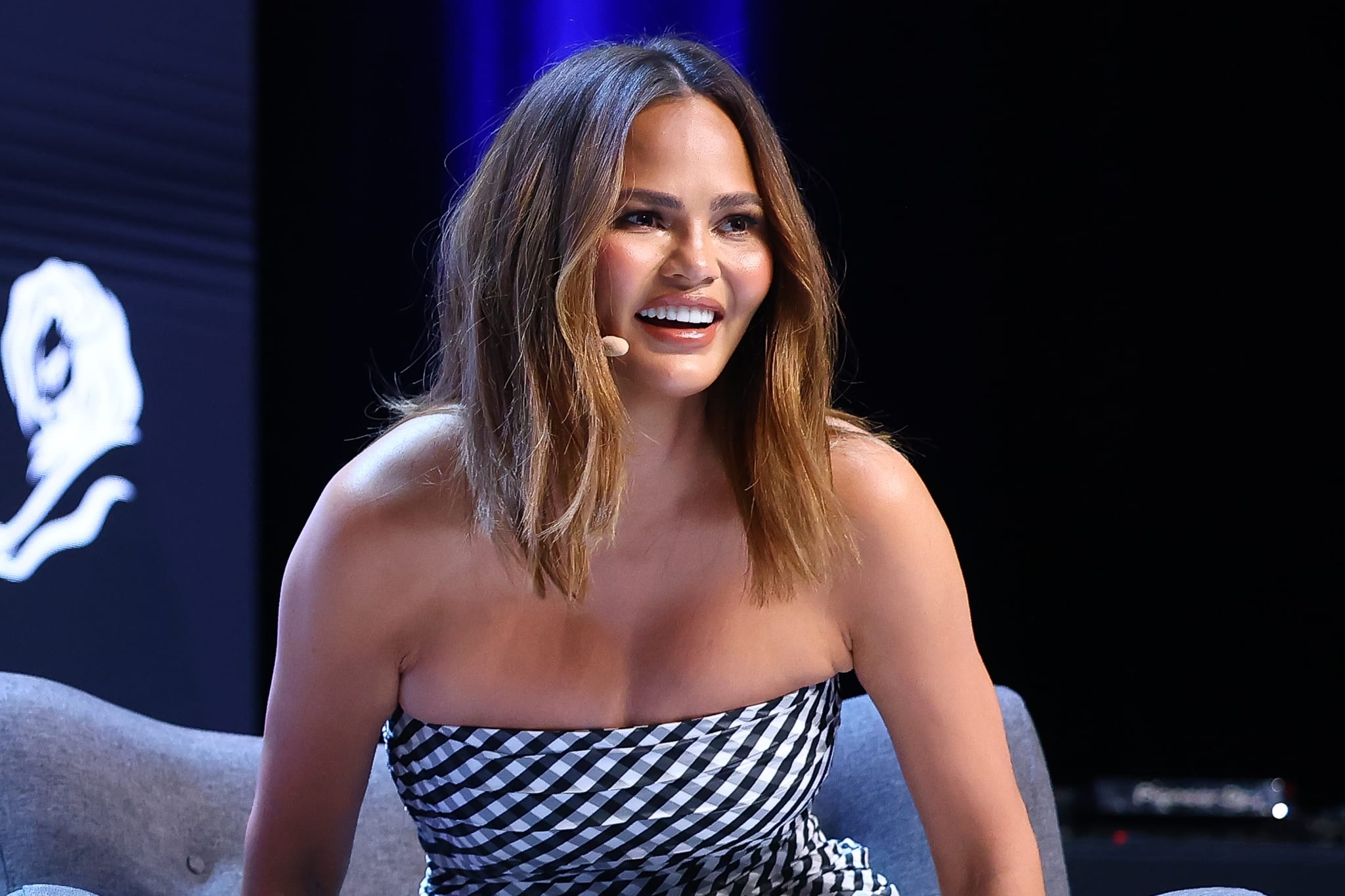 Chrissy Teigen is seated and wearing a strapless checkered dress, smiling while speaking at an event