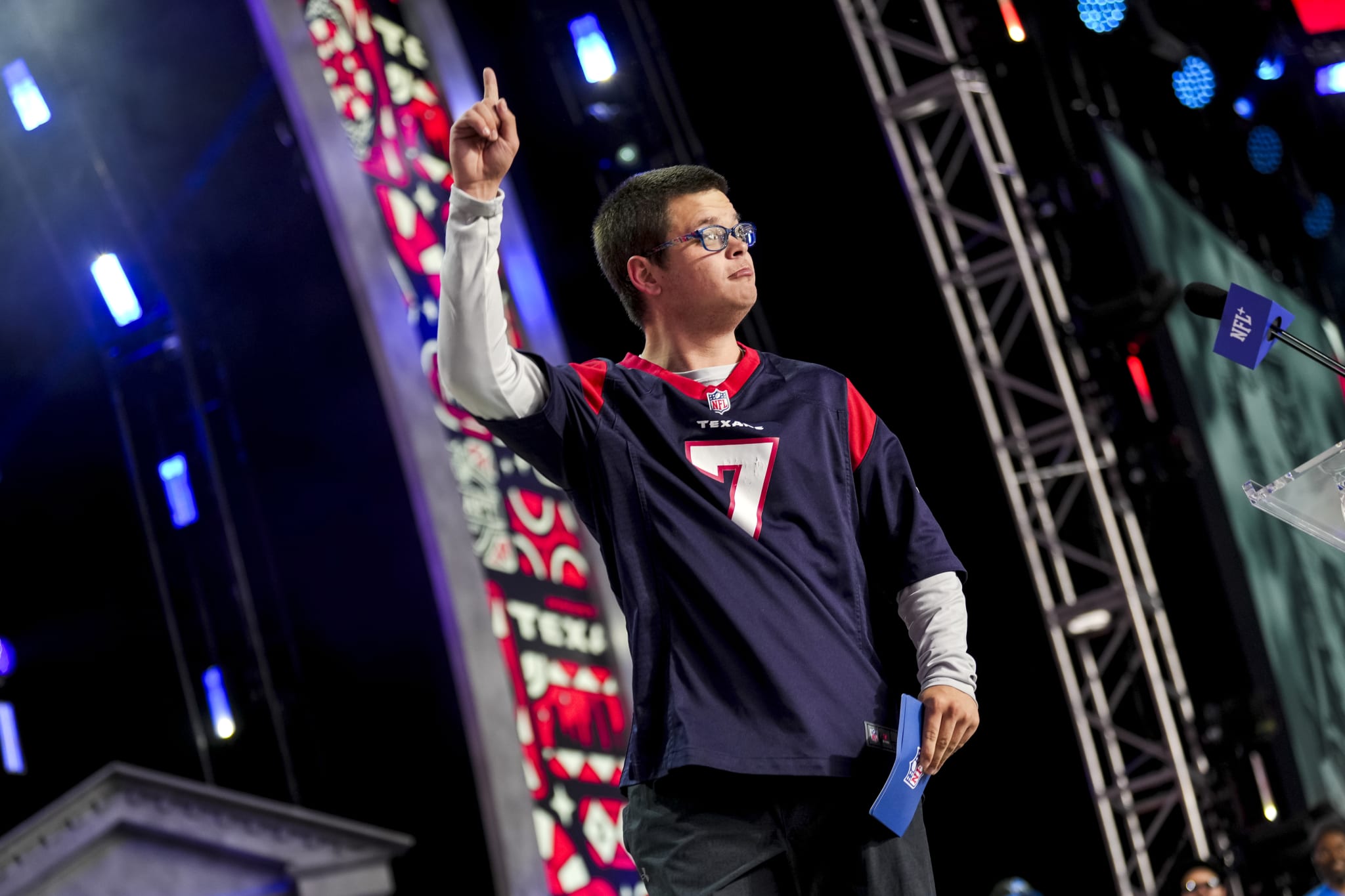 Twitch streamer Sketch reacts after announcnig a pick during round four of the 2024 NFL draft, He is wearing glasses and a sports jersey is on stage, raising one finger, with vibrant lighting and audience in the background.
