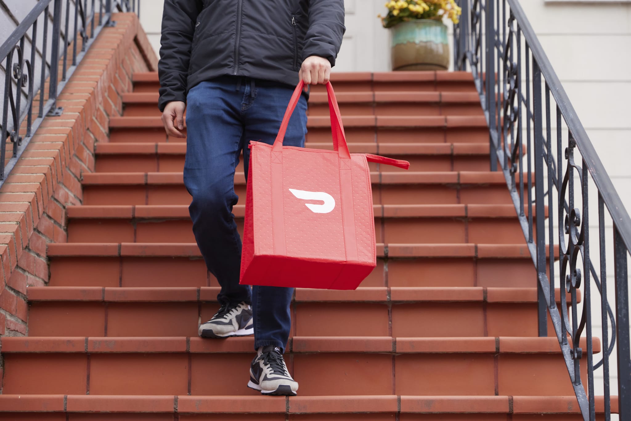 A person in a black jacket and jeans carries a red insulated delivery bag down a set of brick stairs