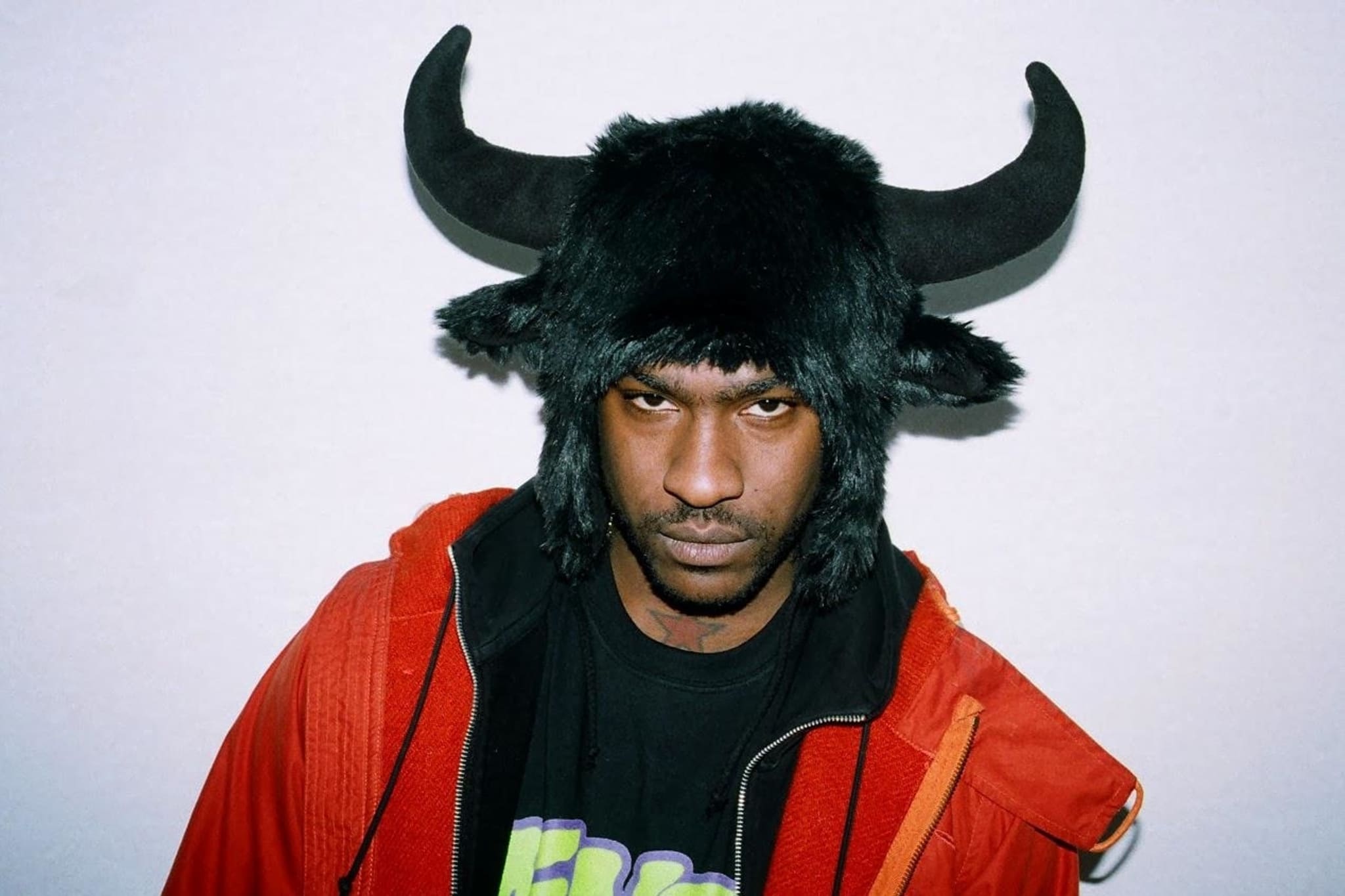 Skepta wears a furry hat with bull horns and a red jacket over a graphic T-shirt