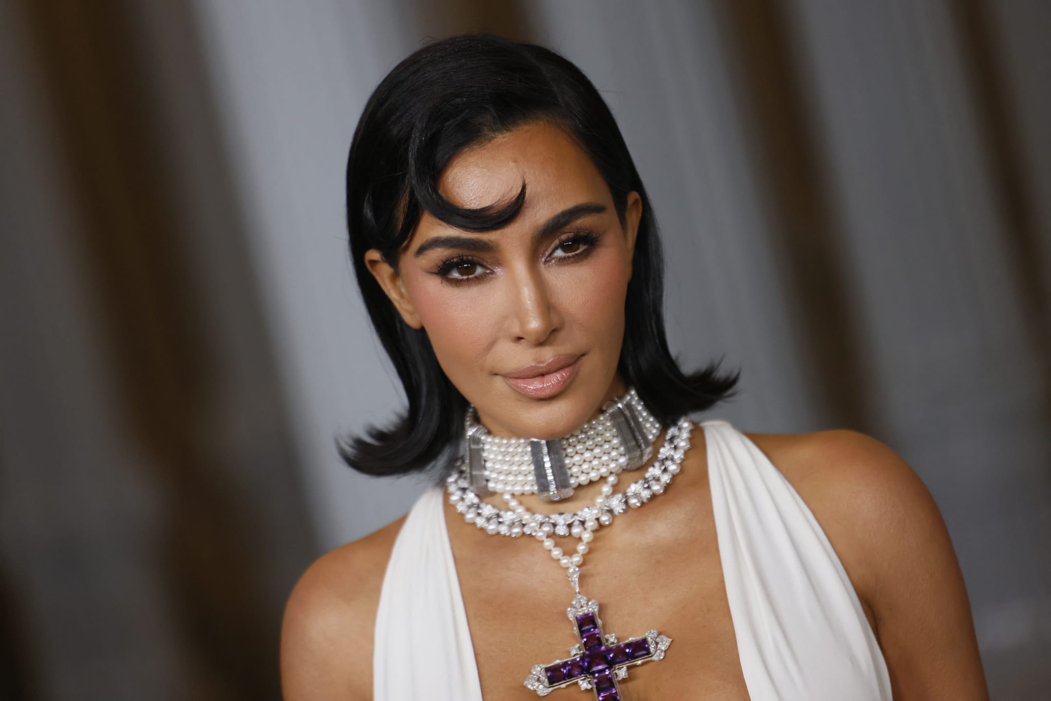Kim Kardashian wearing a white dress, pearl choker, and cross necklace, with sleek black hair styled in waves.