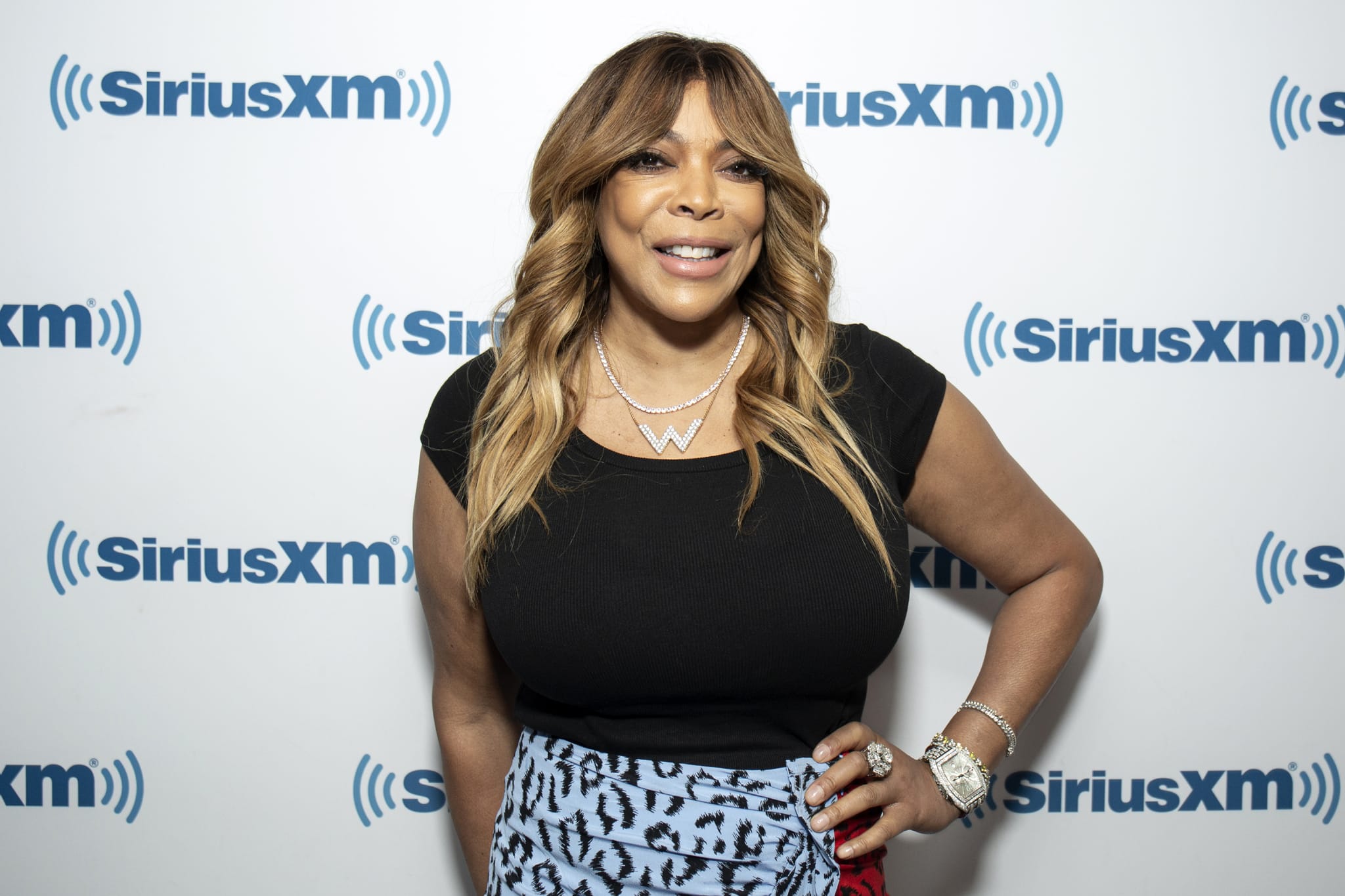 Wendy Williams smiling in front of a backdrop, wearing a black top and patterned skirt, with wavy hair and jewelry.