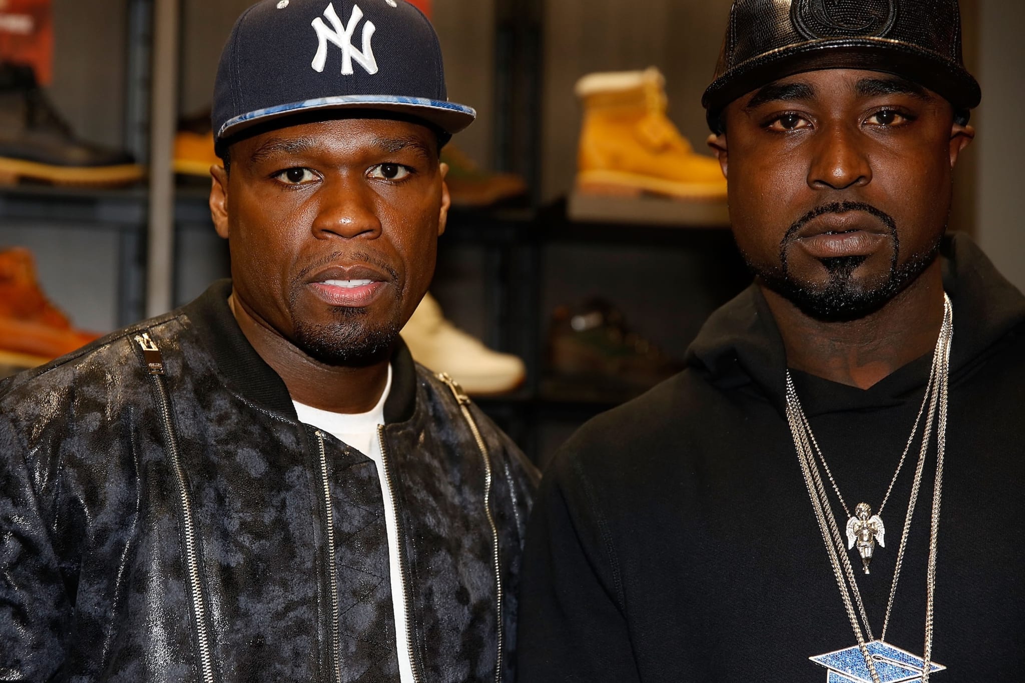 50 Cent and Young Buck pose together, wearing baseball caps and dark jackets, in front of a shoe display.