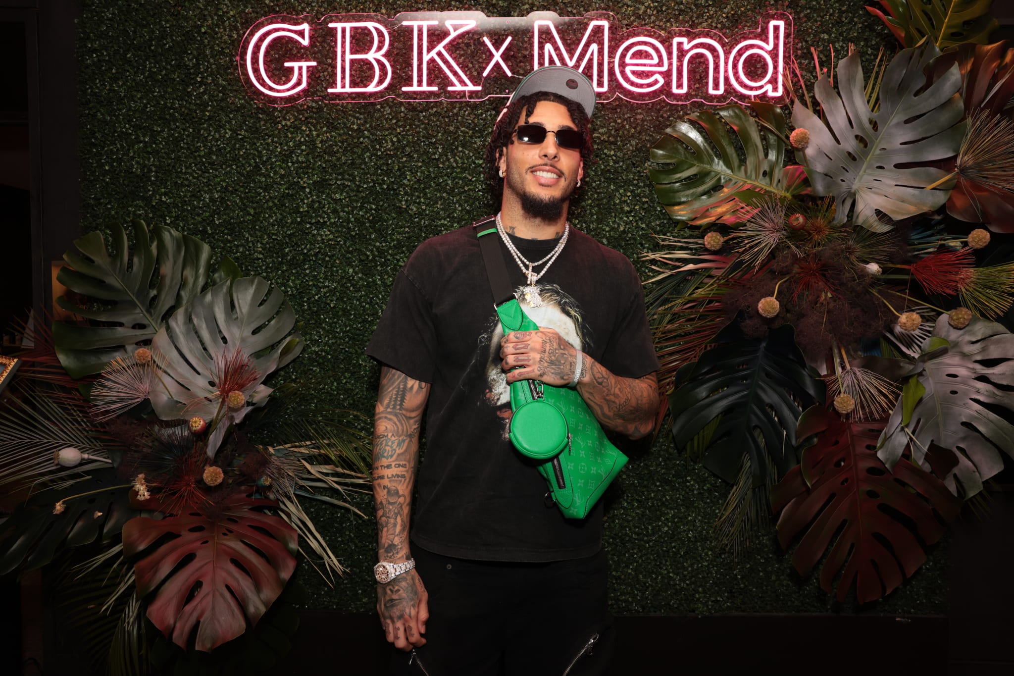 LiAngelo Ball wearing sunglasses and a cap stands in front of a "GBK x Mend" neon sign, surrounded by tropical foliage.