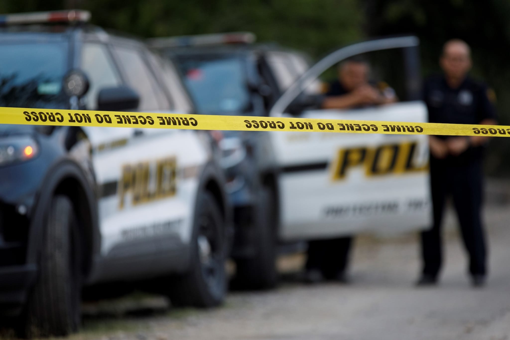 Police vehicles behind yellow crime scene tape with "Do Not Cross" text. Two officers stand near the open door of a patrol car.