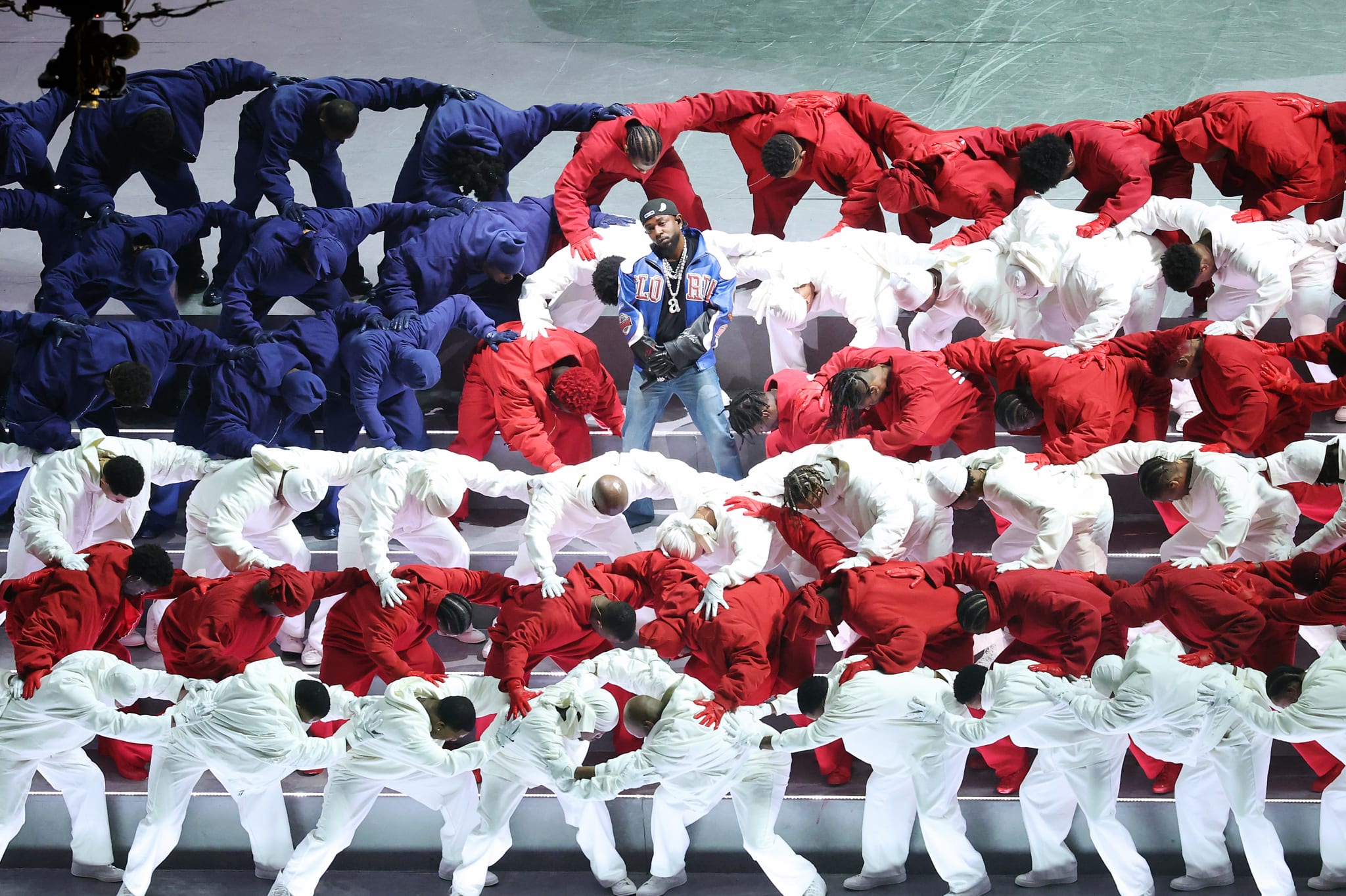 A performer stands surrounded by dancers in blue, red, and white outfits, forming a flag-like pattern on stage.