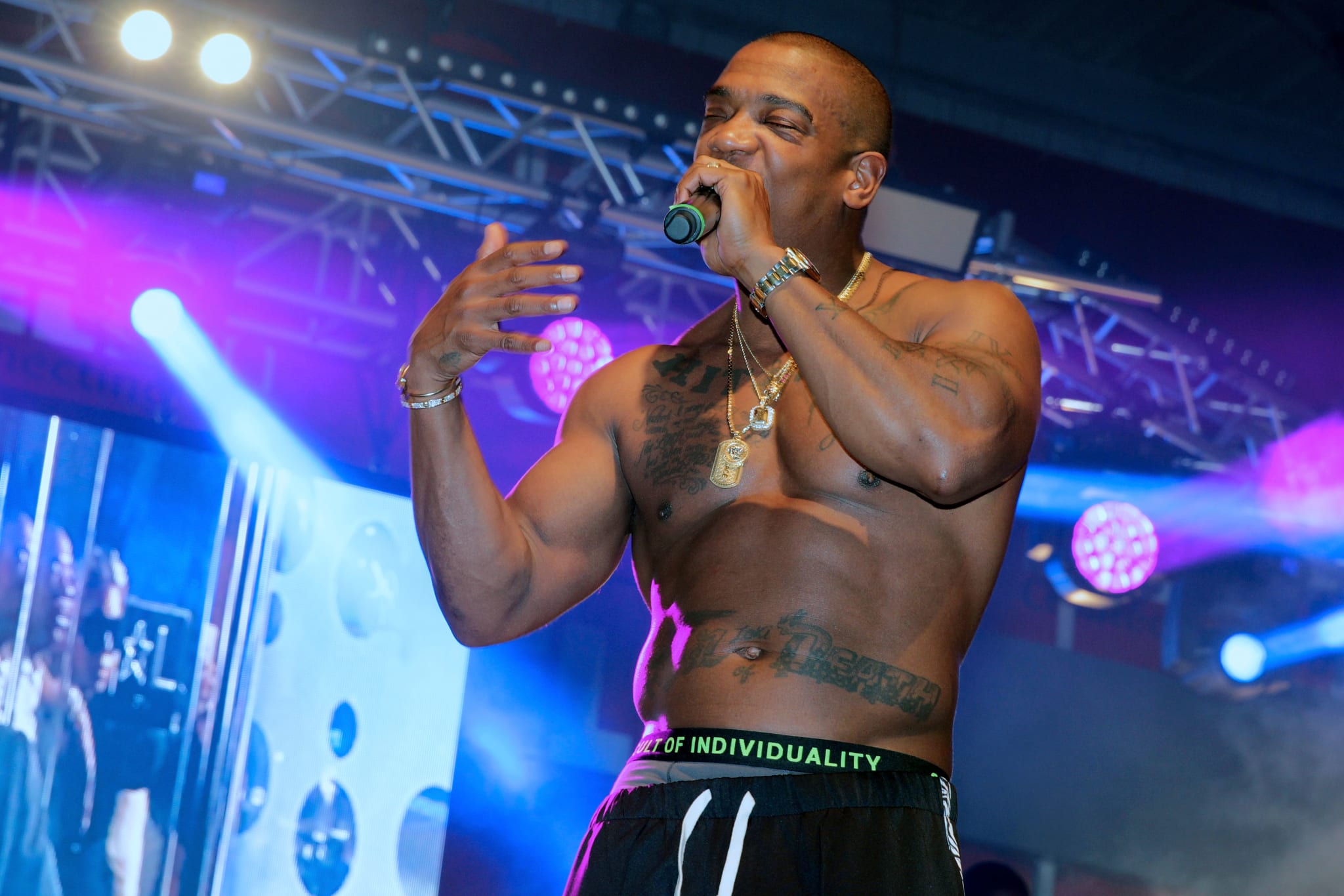 Ja Rule performing shirtless on stage, holding a microphone, with stage lights and equipment in the background.