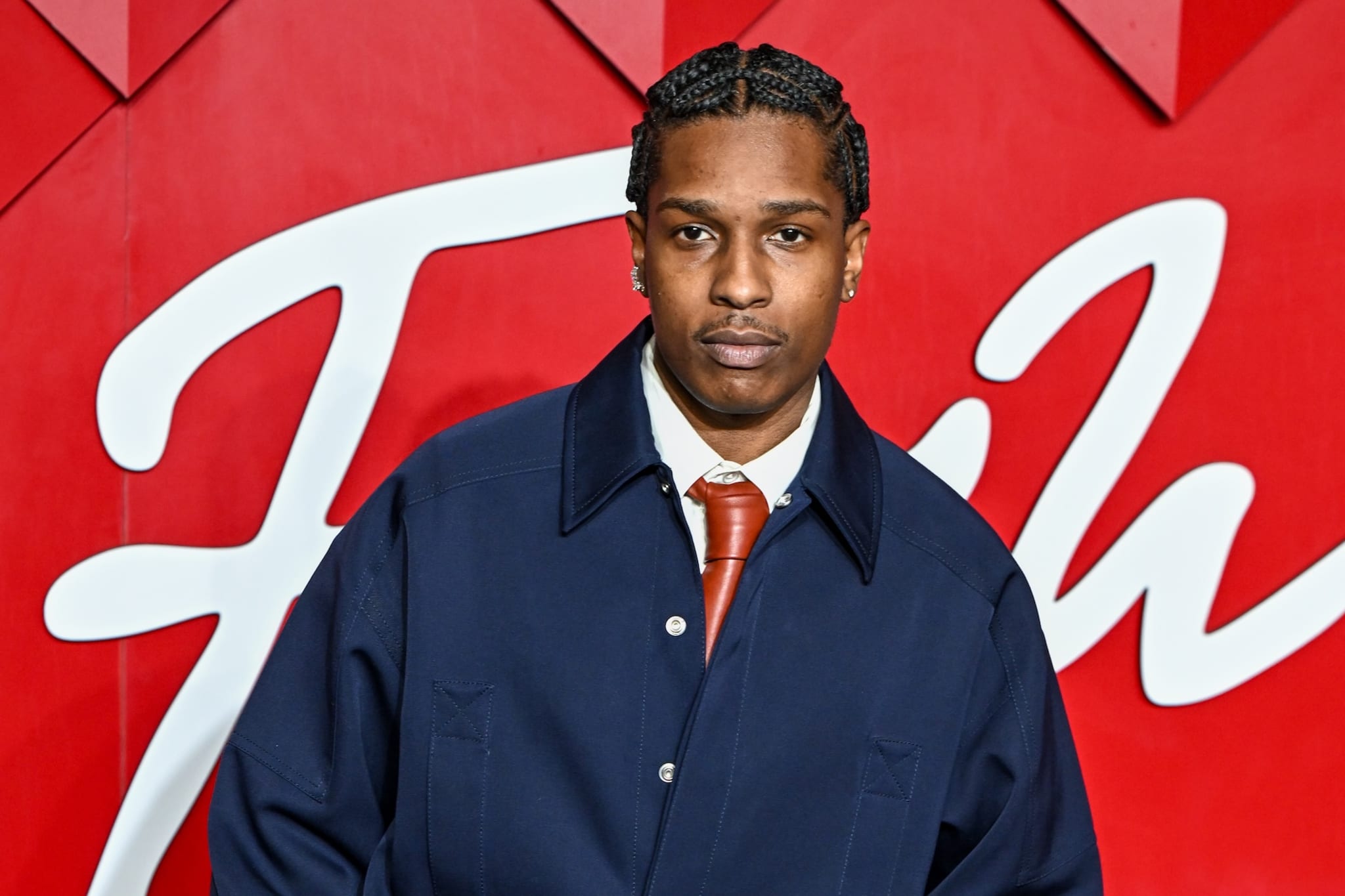 A$AP Rocky in a navy jacket and orange tie, standing in front of a red background with white script.